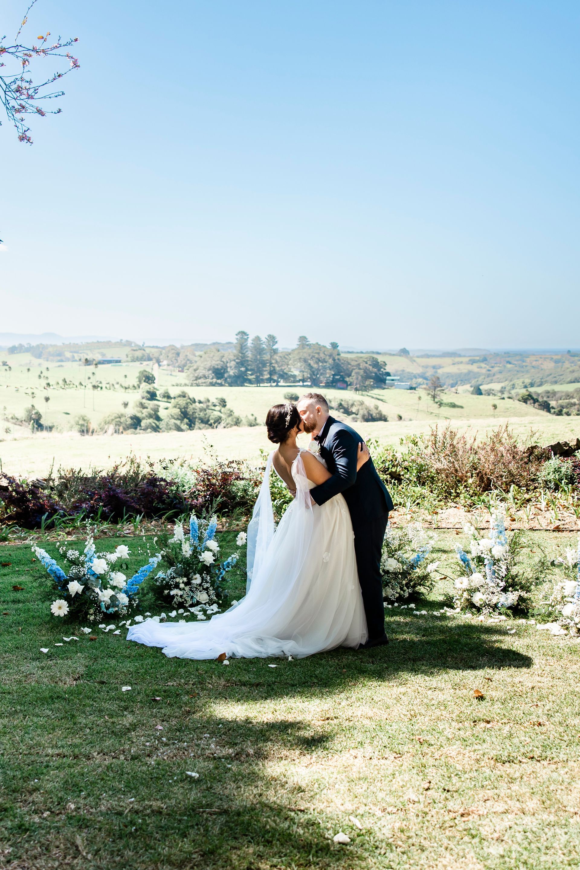 Bride and groom kissing in a garden with white flowers and a scenic countryside backdrop