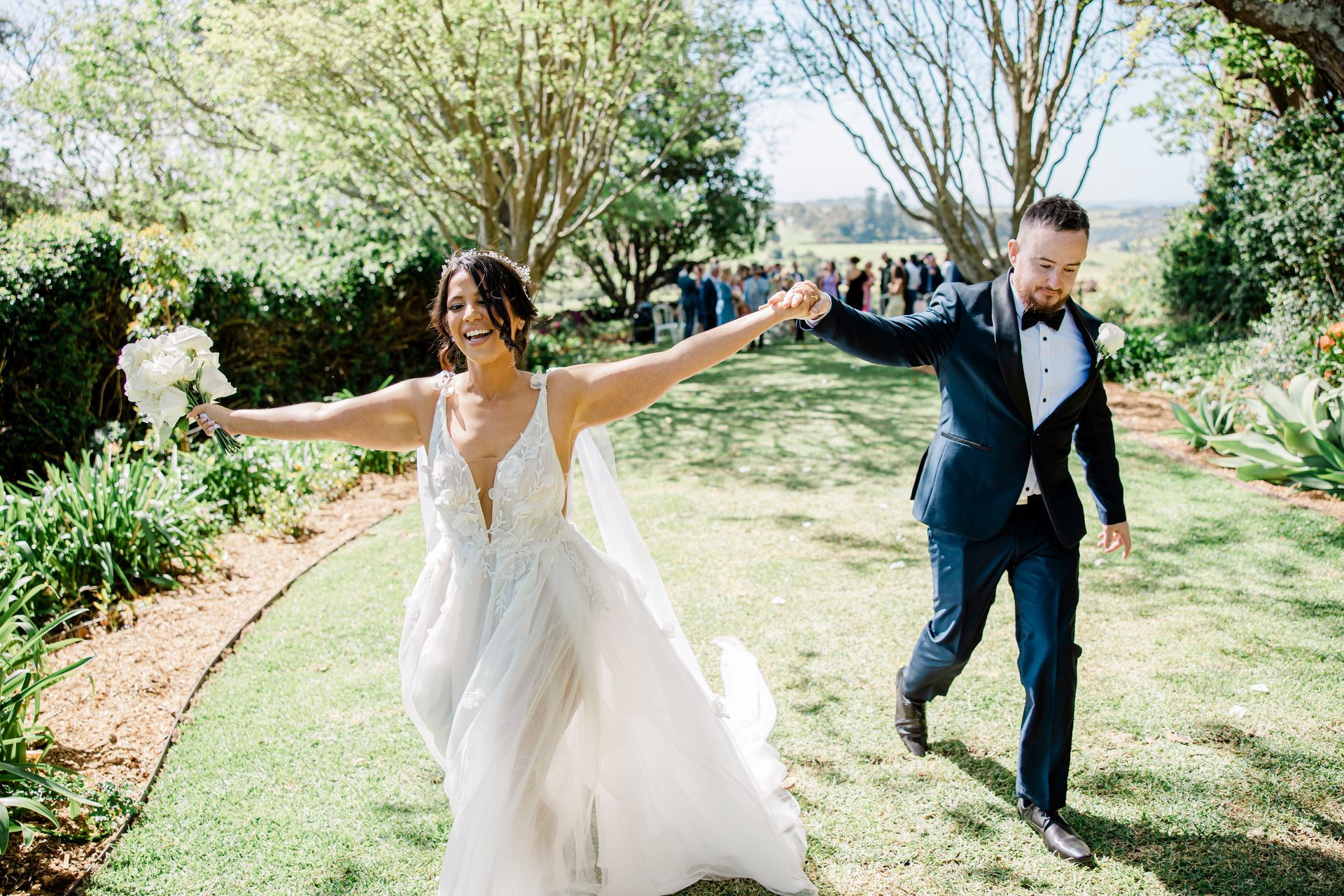 Bride and groom run hand-in-hand down a garden path, with arms outstretched in a bright outdoor setting.
