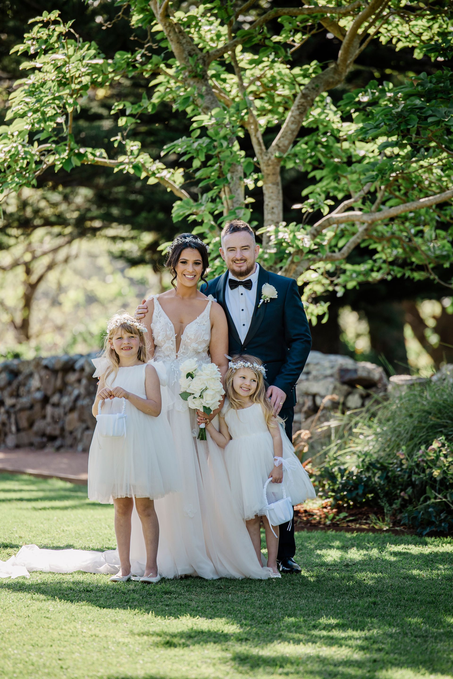 Wedding couple with three children posing outdoors under a tree, bride holding bouquet in white gown