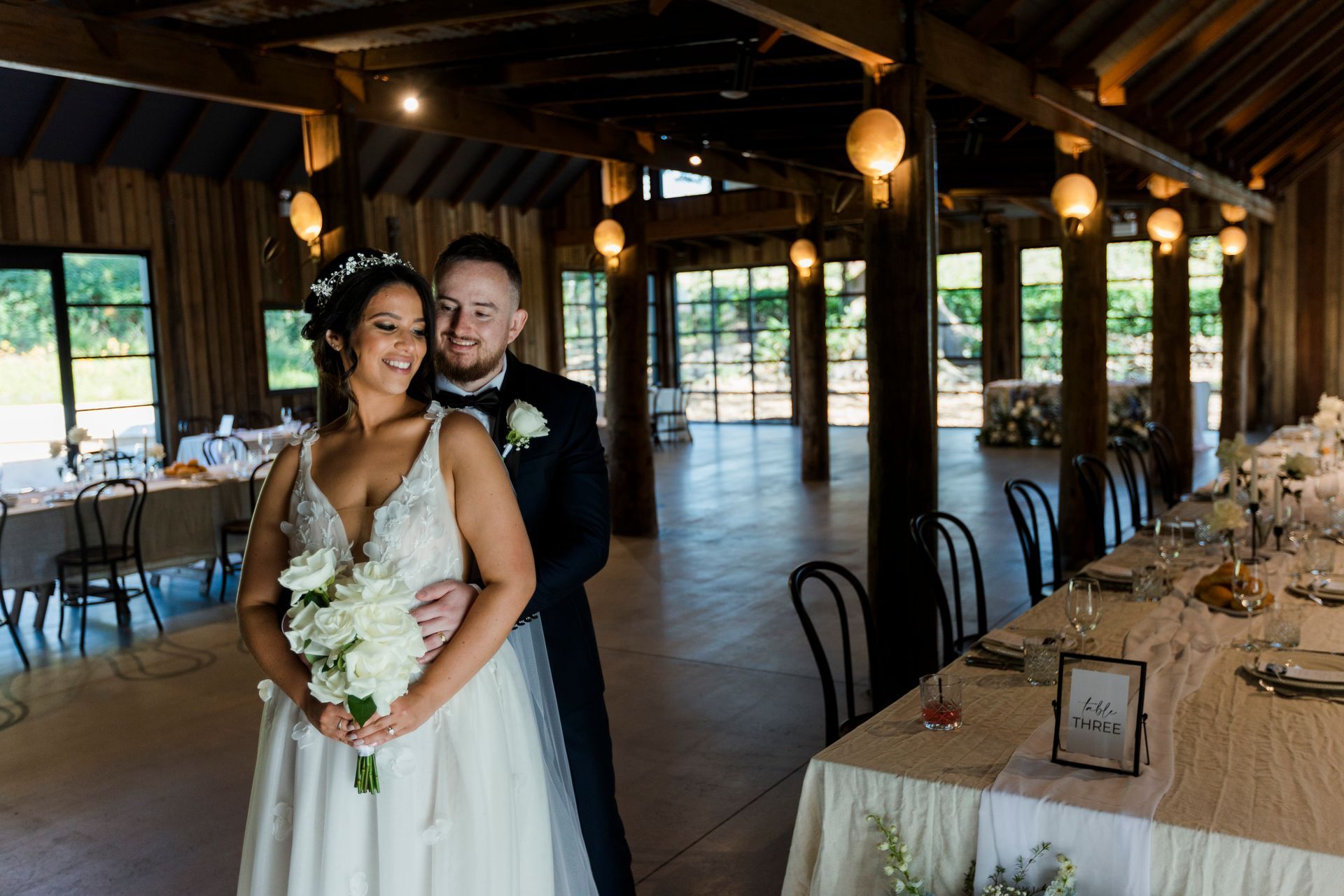 Bride and groom embrace in a rustic reception hall with warm lights and long banquet tables