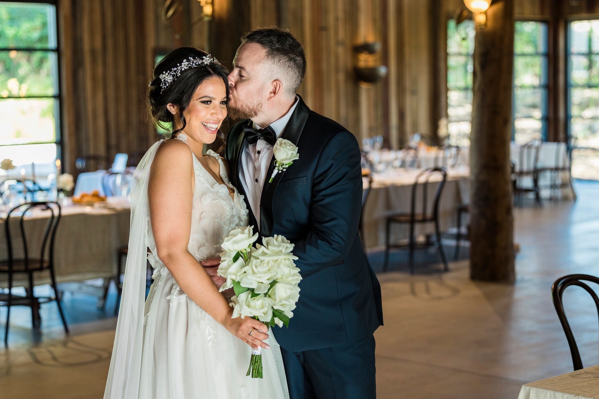 Bride and groom sharing a kiss in a rustic wedding venue, bride holding a white bouquet