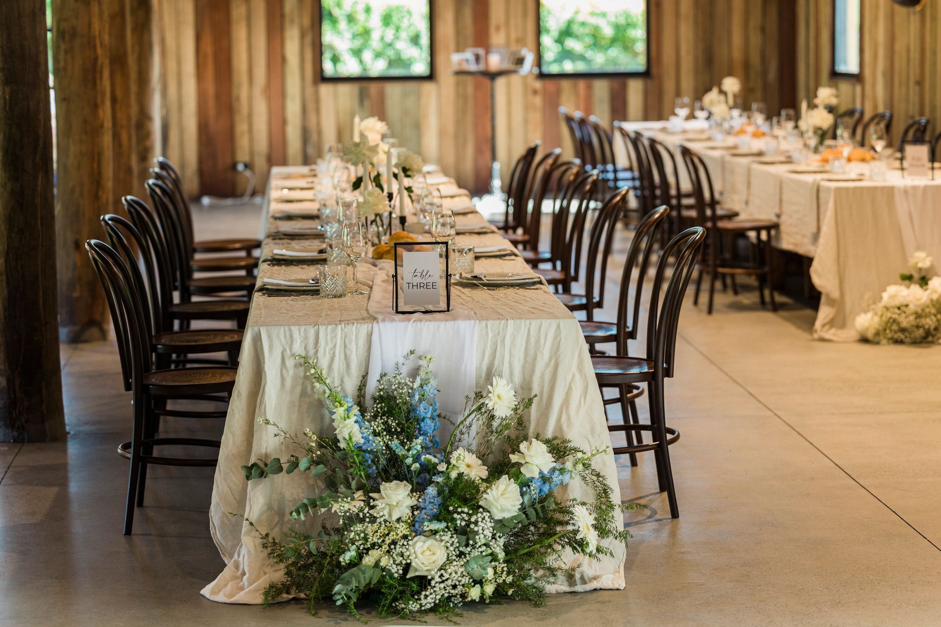 Elegant banquet tables in a rustic hall, decorated with white linens and blue floral centerpieces.