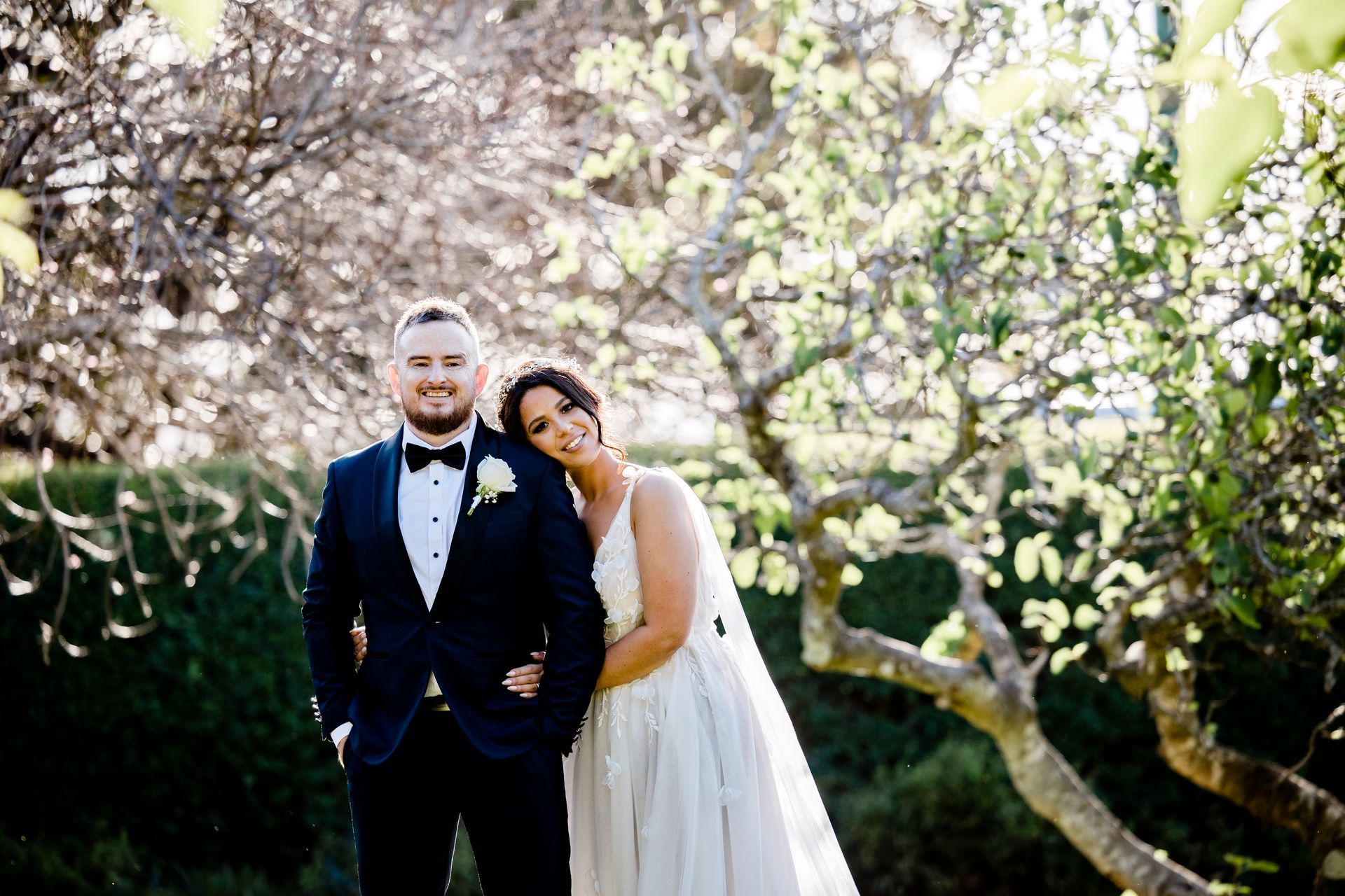 Couple in wedding attire posing outdoors beneath blooming trees