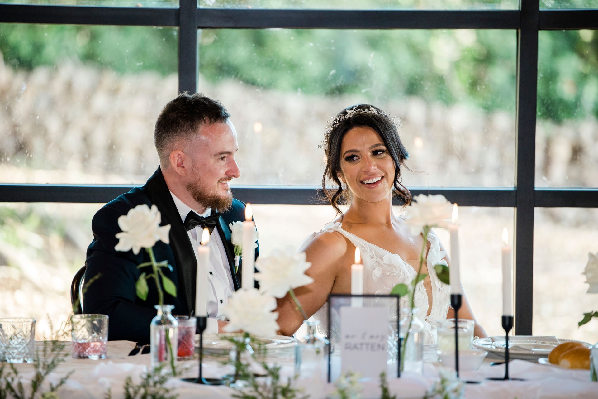 Bride and groom smiling at a wedding reception table with white flowers and candles