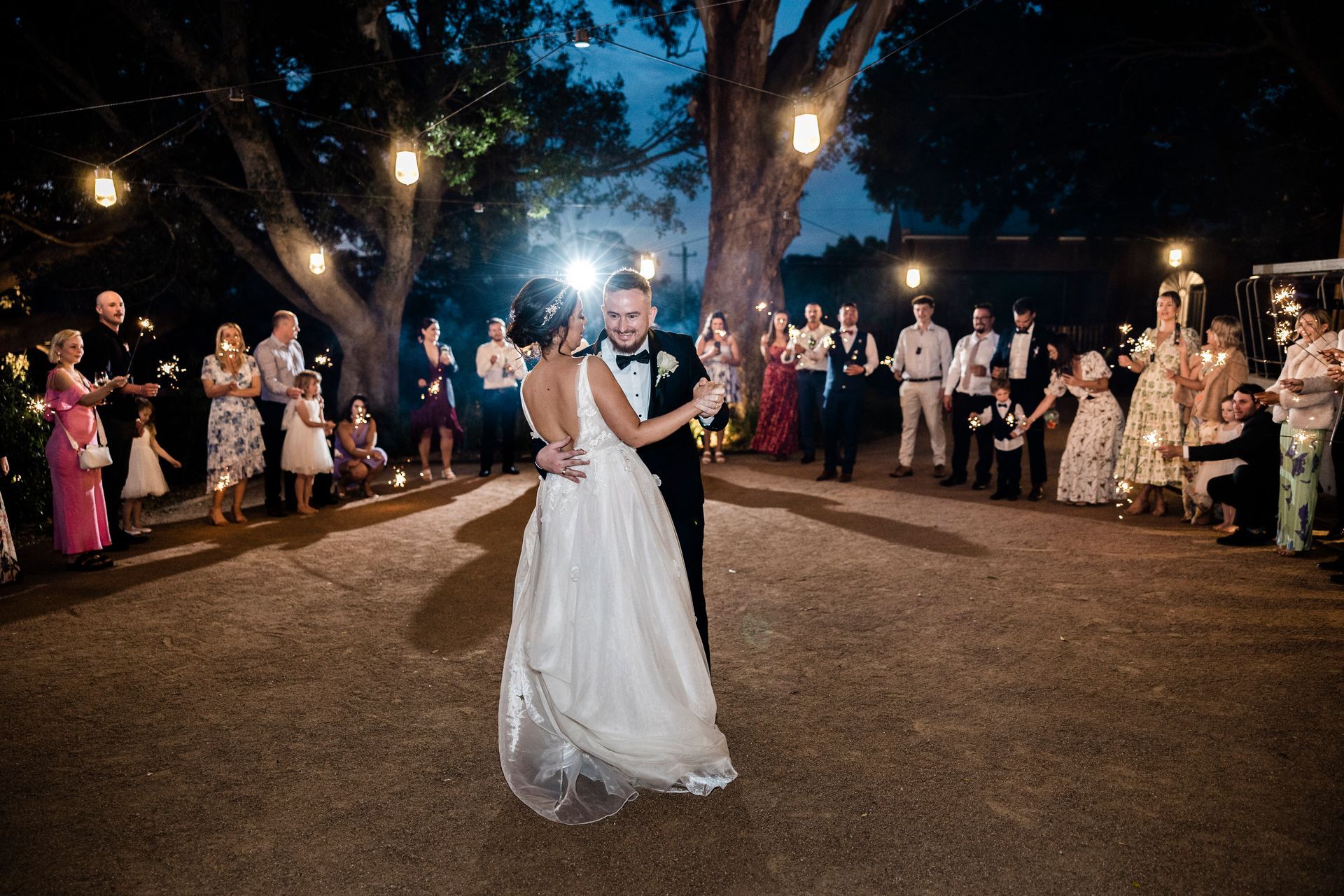 Bride and groom dancing in a lit outdoor wedding reception, guests lined along the path at night.