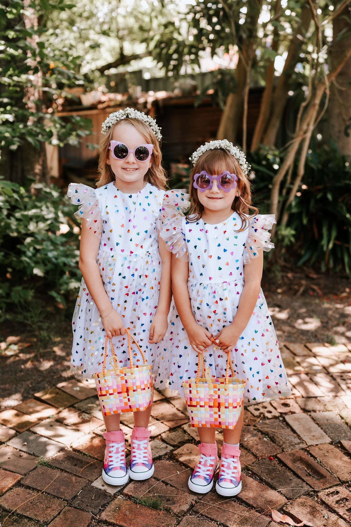 Two children in matching white dresses and sunglasses standing on a brick path outdoors