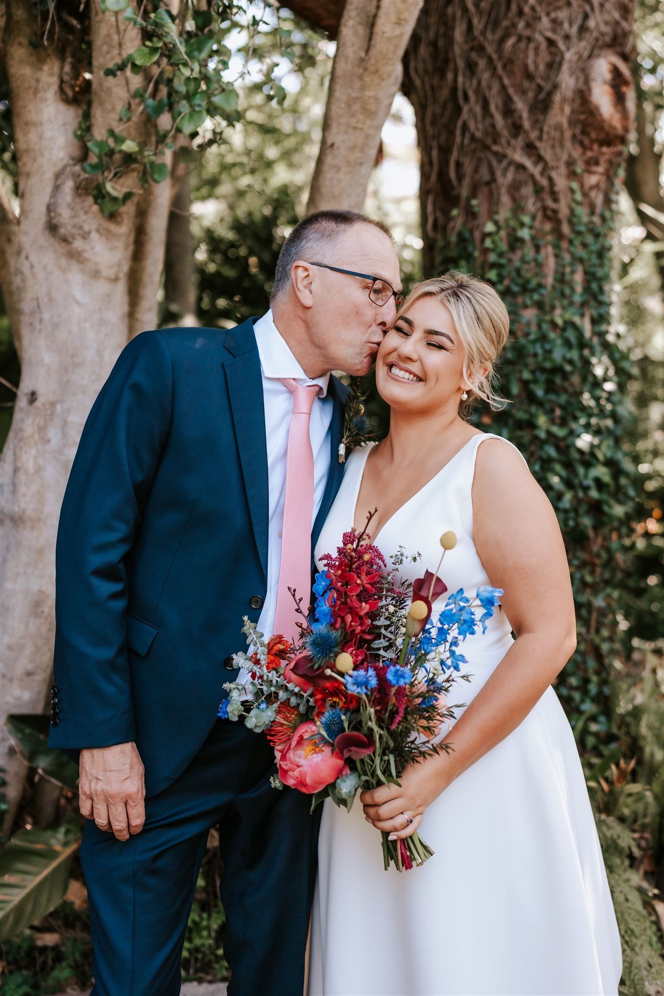 Couple posing outdoors, bride in white dress holding colorful bouquet while groom kisses her cheek.
