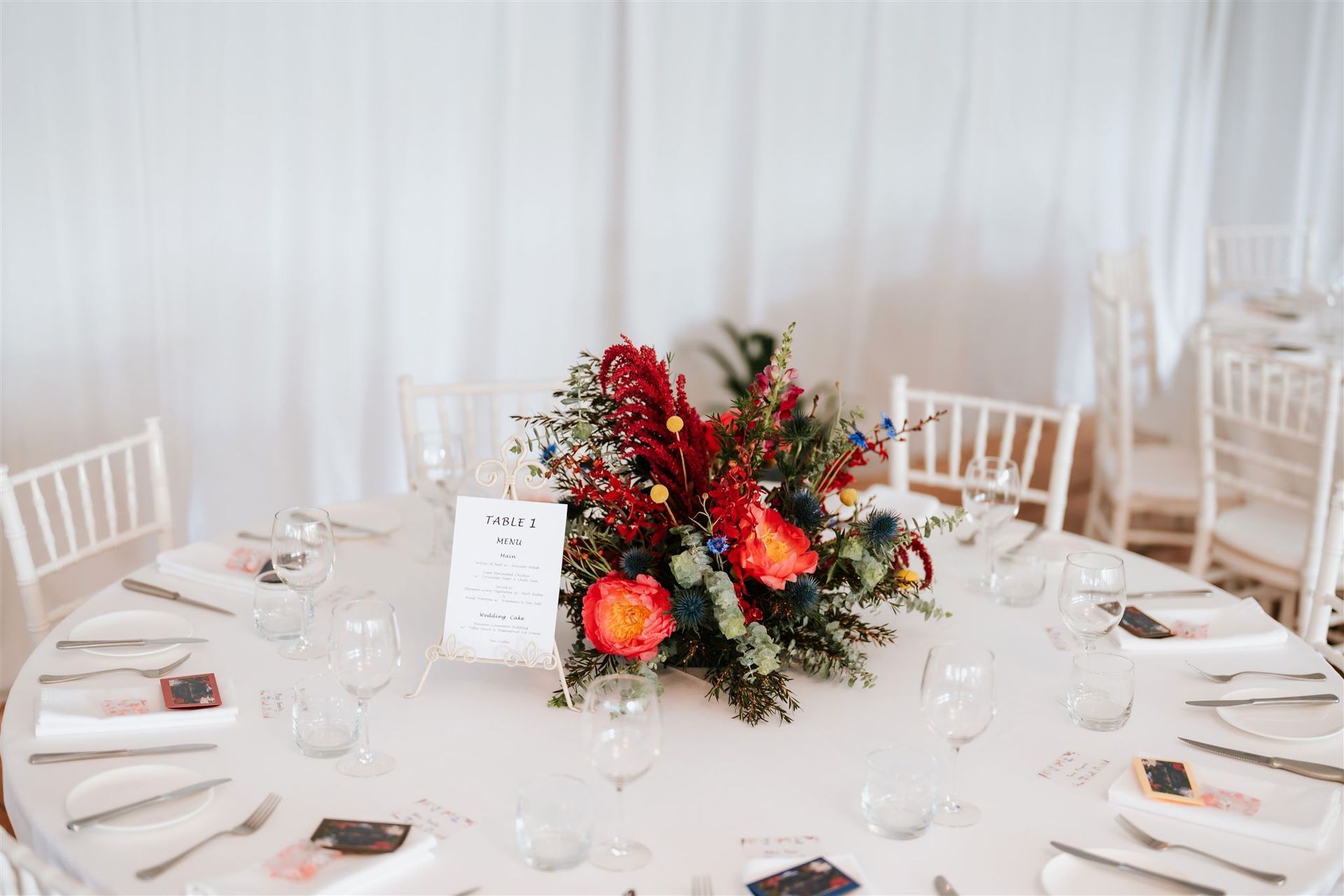 Elegant white-table wedding reception with a red and orange floral centerpiece and place settings