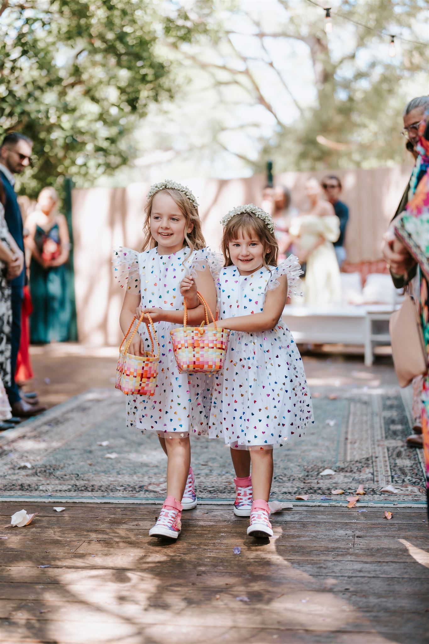 Two children in matching white polka-dot dresses walk down an outdoor aisle holding flower baskets.