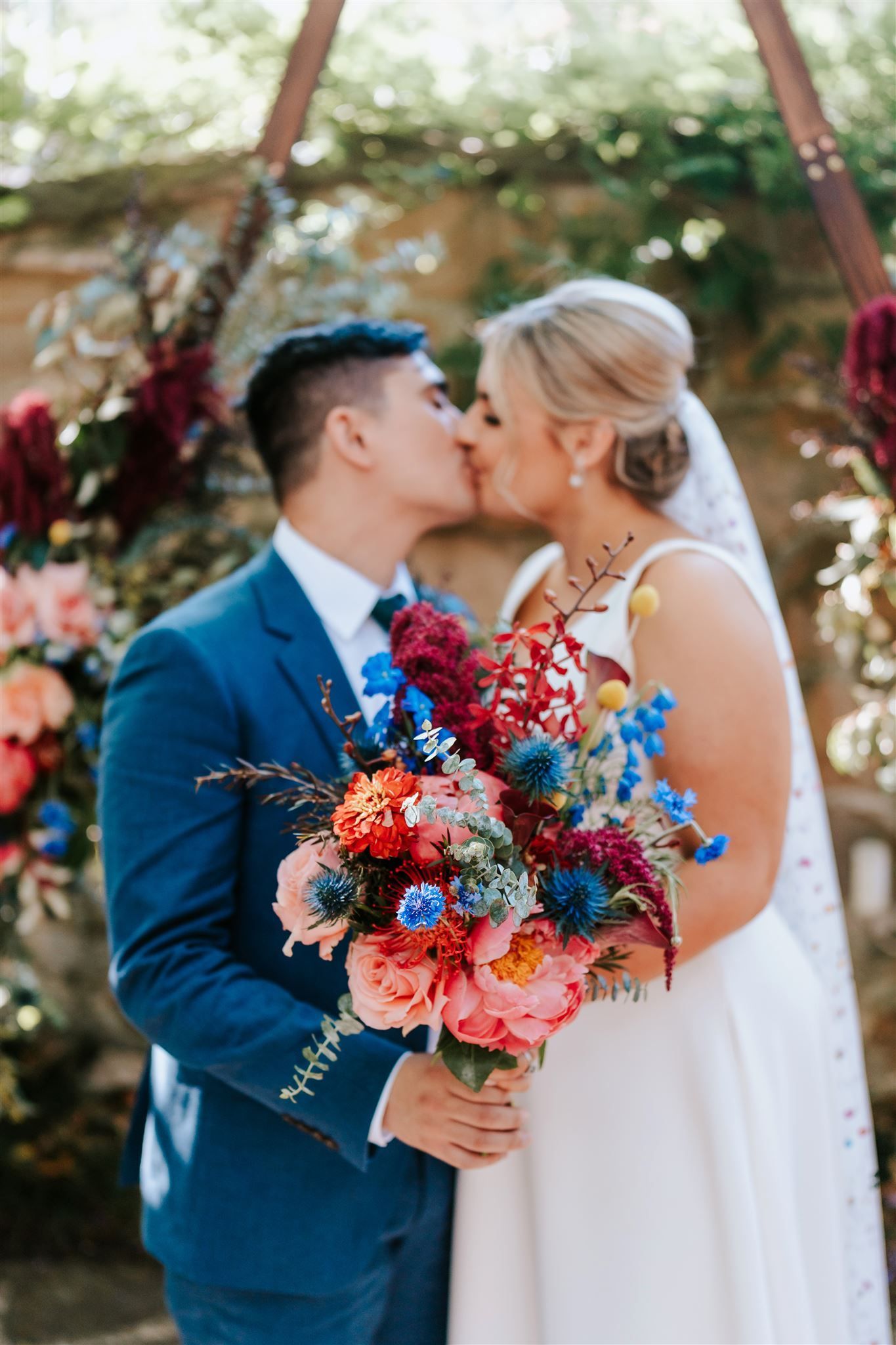 Bride and groom kissing, holding a vibrant bouquet in a floral outdoor wedding setting