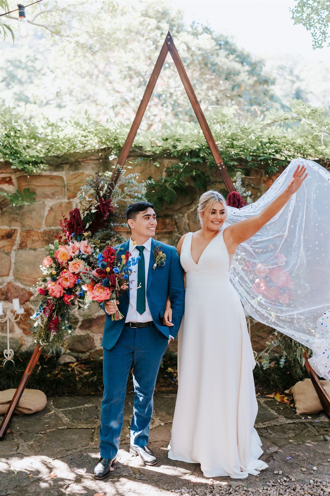 Wedding couple posing outdoors under floral arch, bride lifting veil beside groom in blue suit