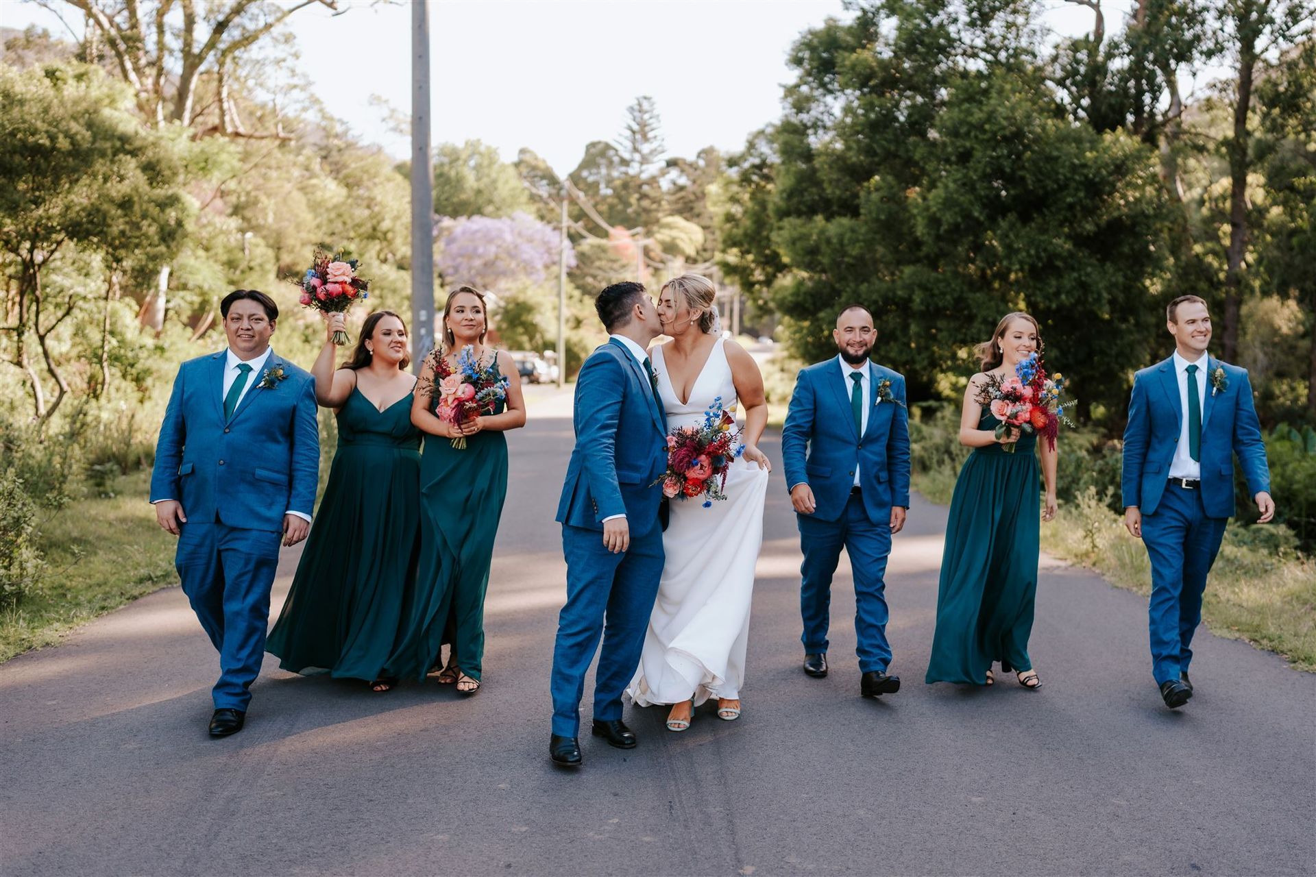 Wedding party walking down a tree-lined road, with the bride in white and attendants in teal and blue suits and dresses