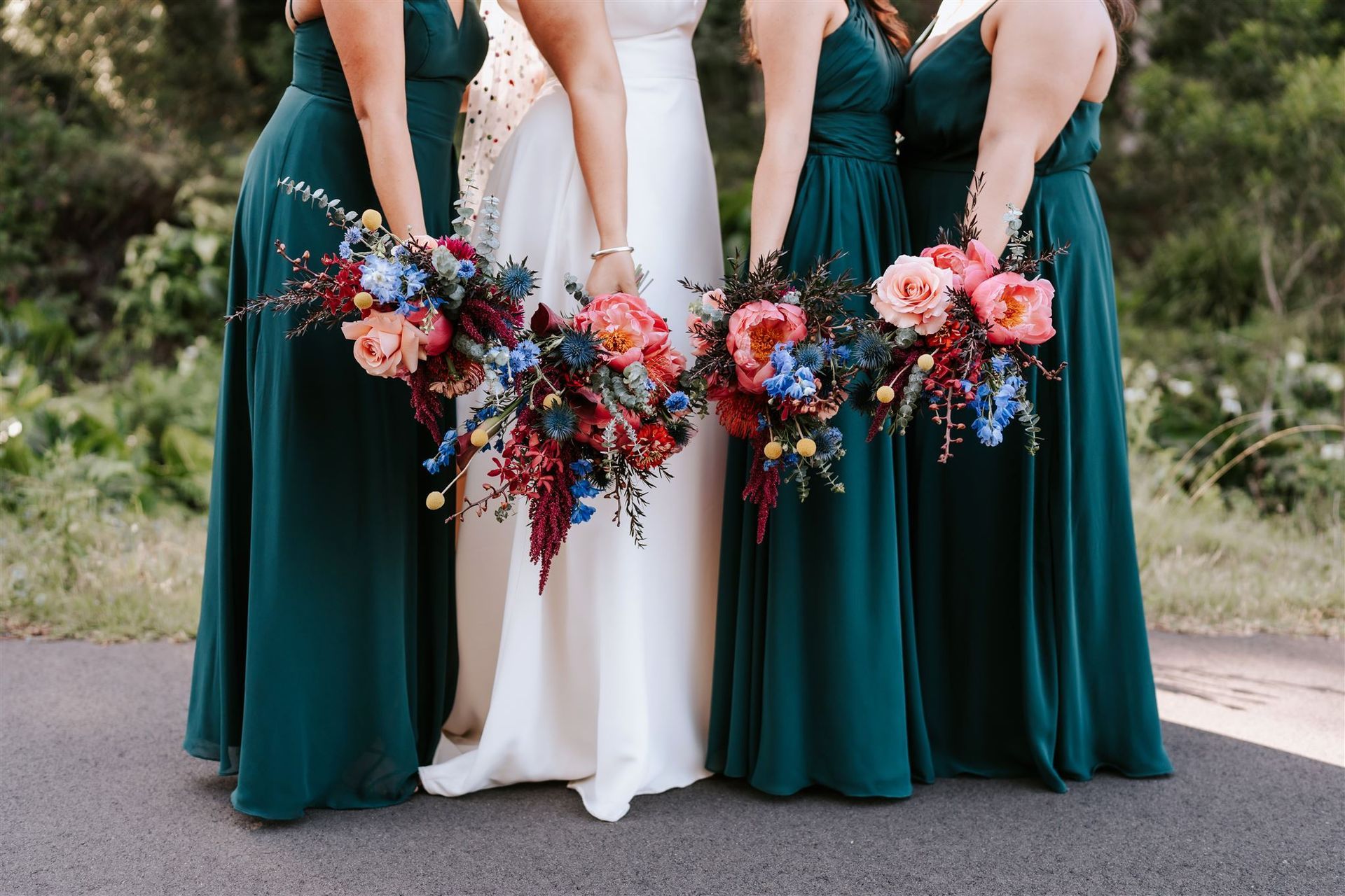 Three bridesmaids in teal dresses and a bride in white holding colorful bouquets outdoors