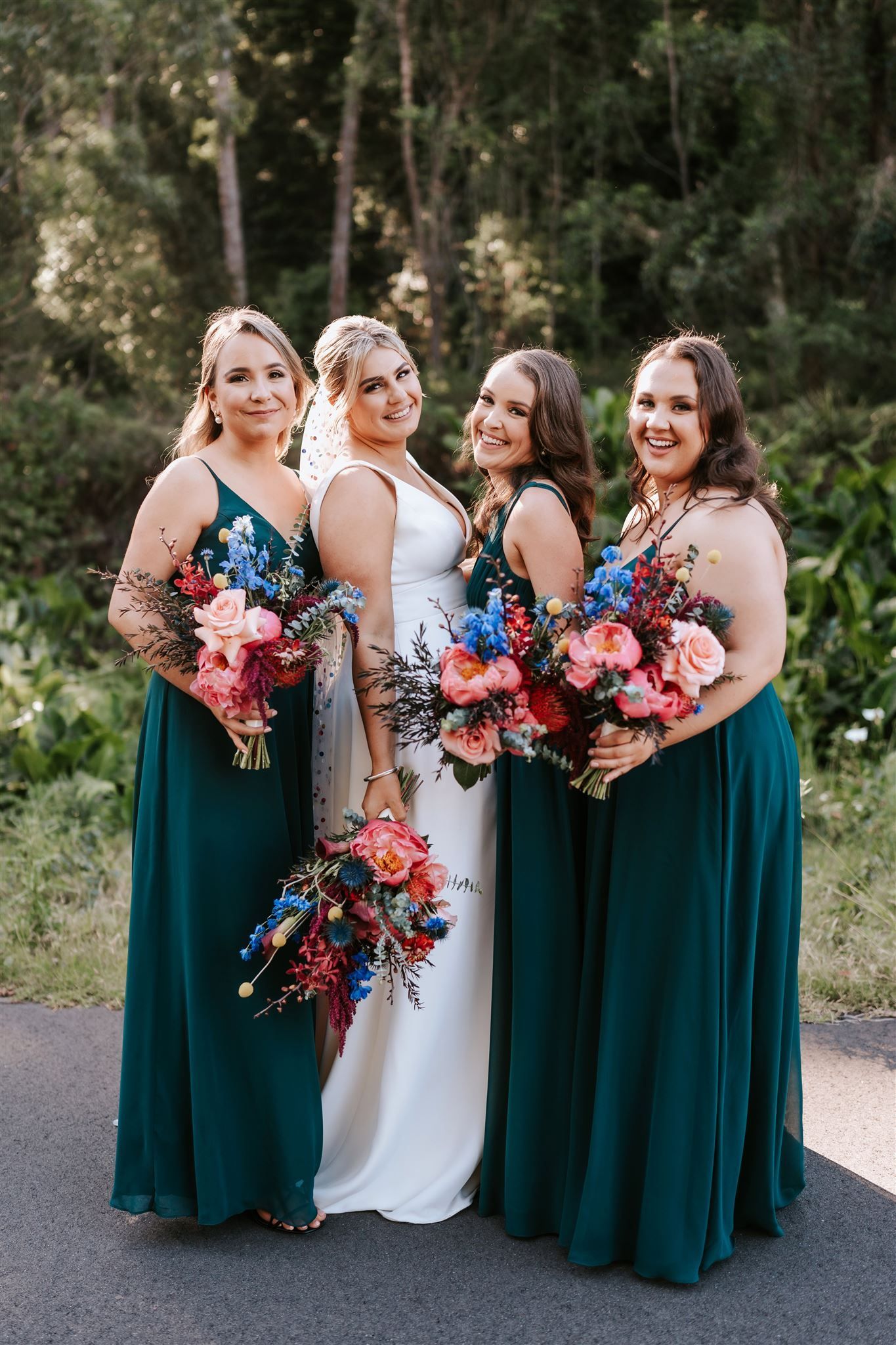 Four people in formal dresses holding colorful bouquets outdoors in a wooded setting.