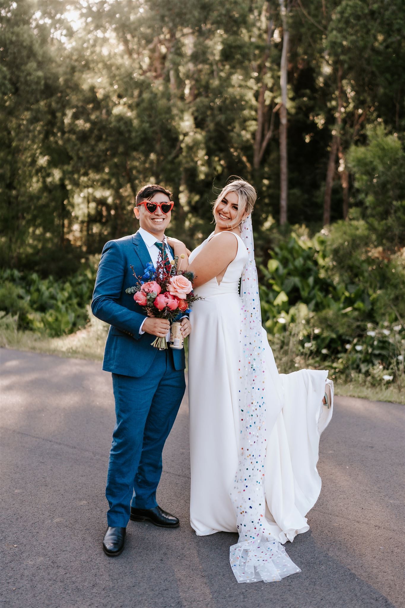 Wedding couple standing outdoors, holding a bouquet, with greenery in the background.