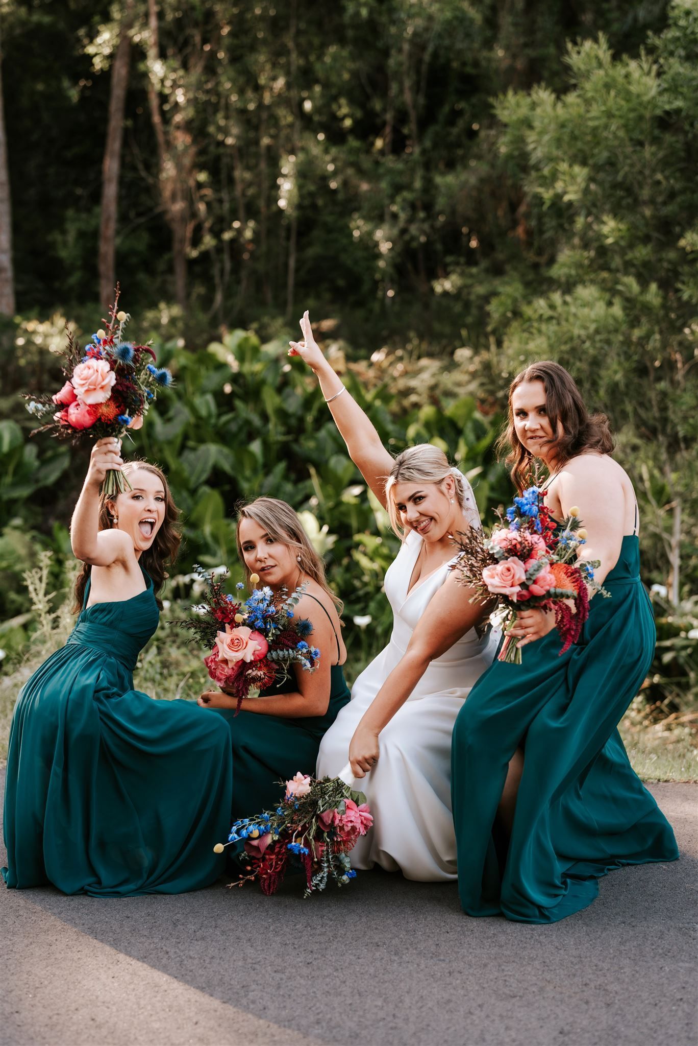 Four women in teal and white dresses pose on a road holding colorful bouquets in a wooded setting