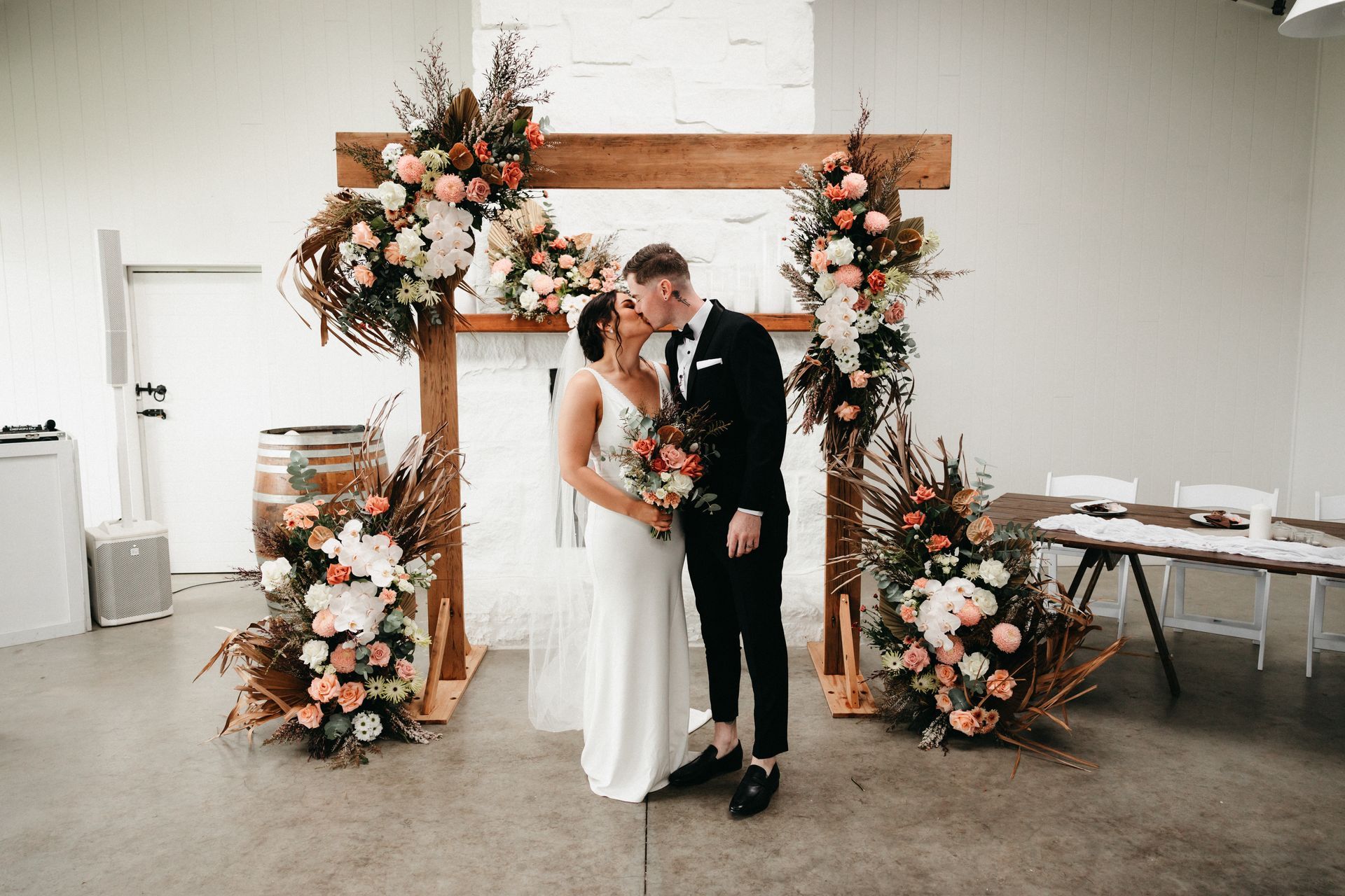 Bride and groom kiss under a floral wedding arch in a bright indoor venue