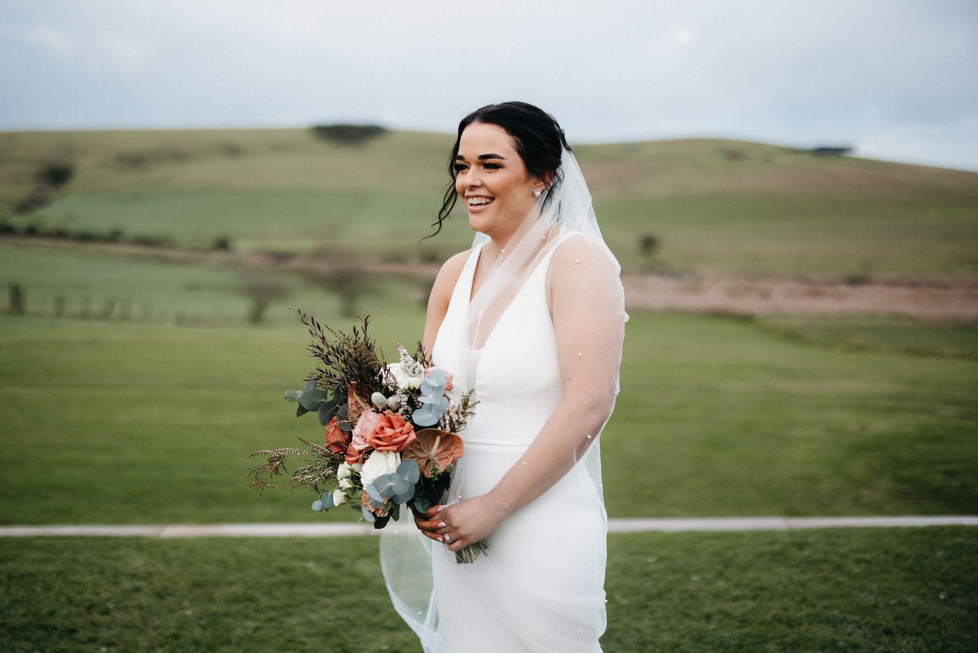 Bride in a white dress holding a bouquet, smiling outdoors in a grassy field.