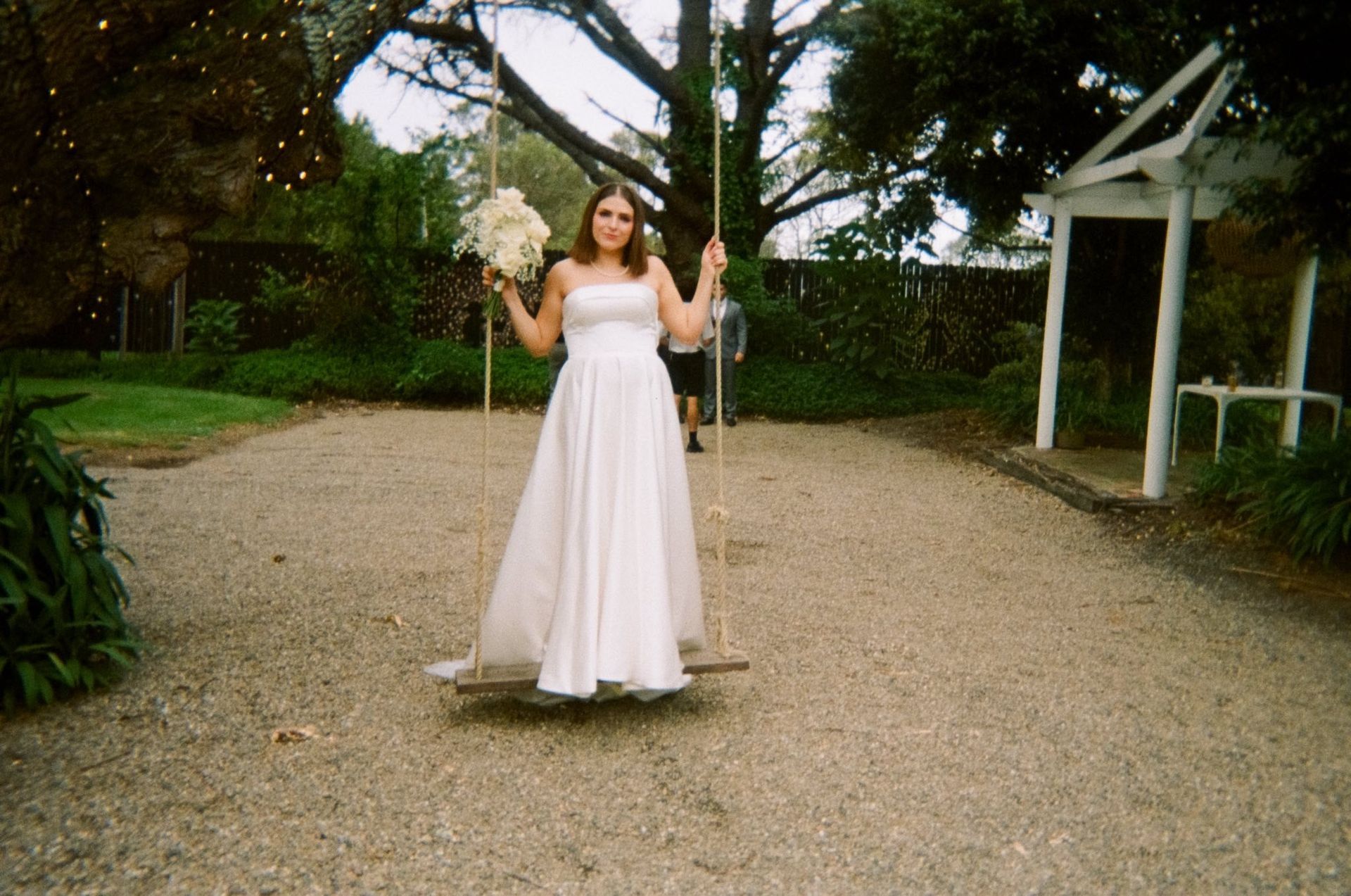Woman in a white dress standing on a swing in a garden path, holding the ropes.