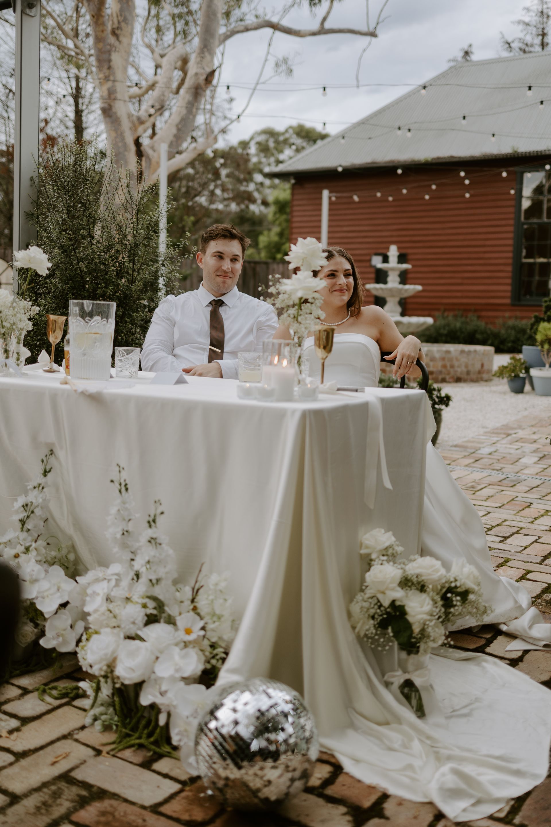 Wedding couple seated at a white floral table outdoors in a cobblestone courtyard