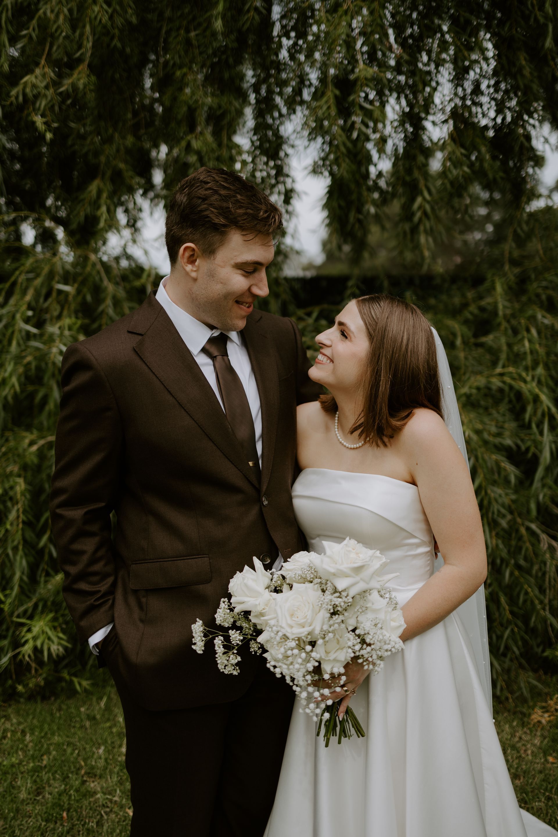Bride and groom smiling at each other outdoors, bride holding a white bouquet in a white dress and groom in a dark suit