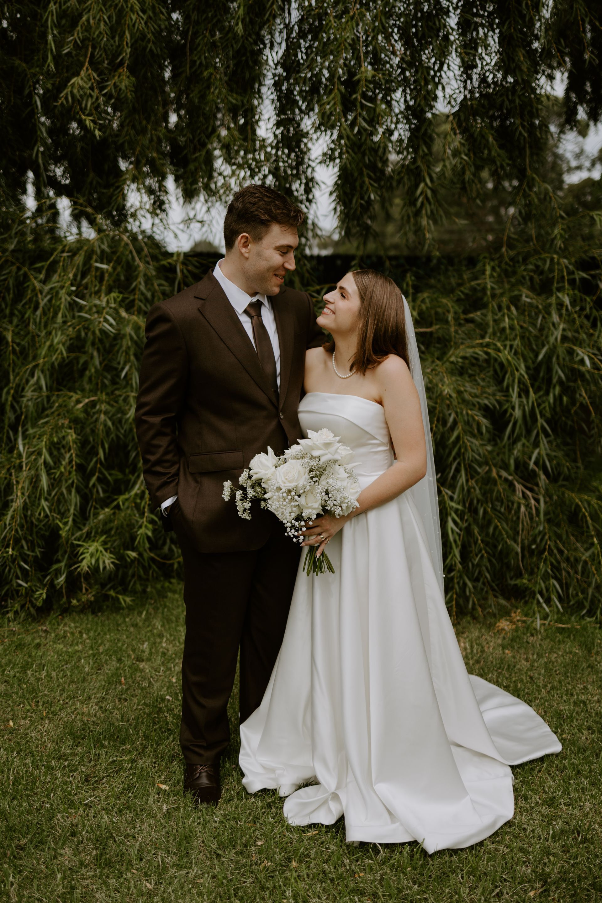 Bride and groom standing together outdoors, smiling and holding a white bouquet