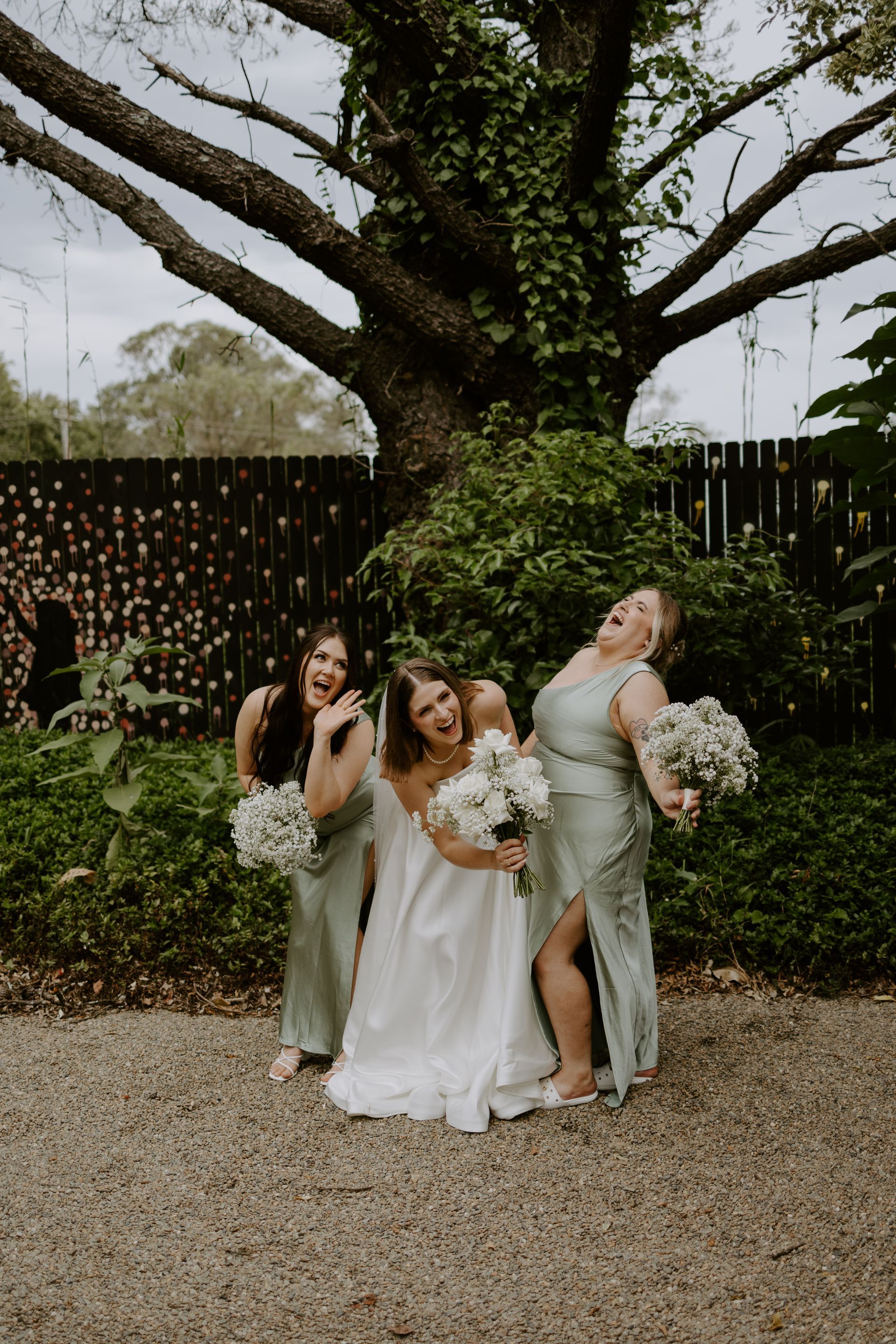 Three women posing with bouquets under a large tree in a garden setting