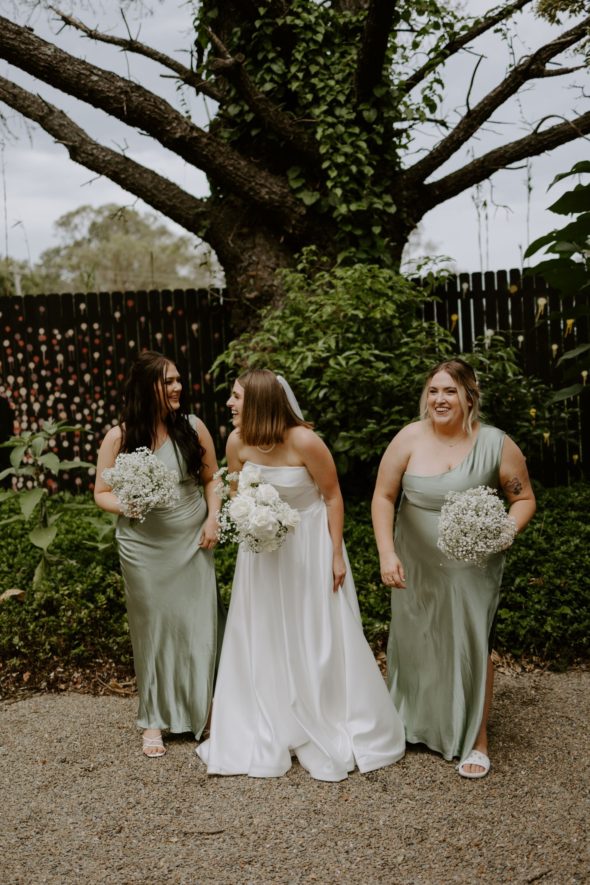 Three women in formal dresses holding white bouquets pose outdoors under a large tree.
