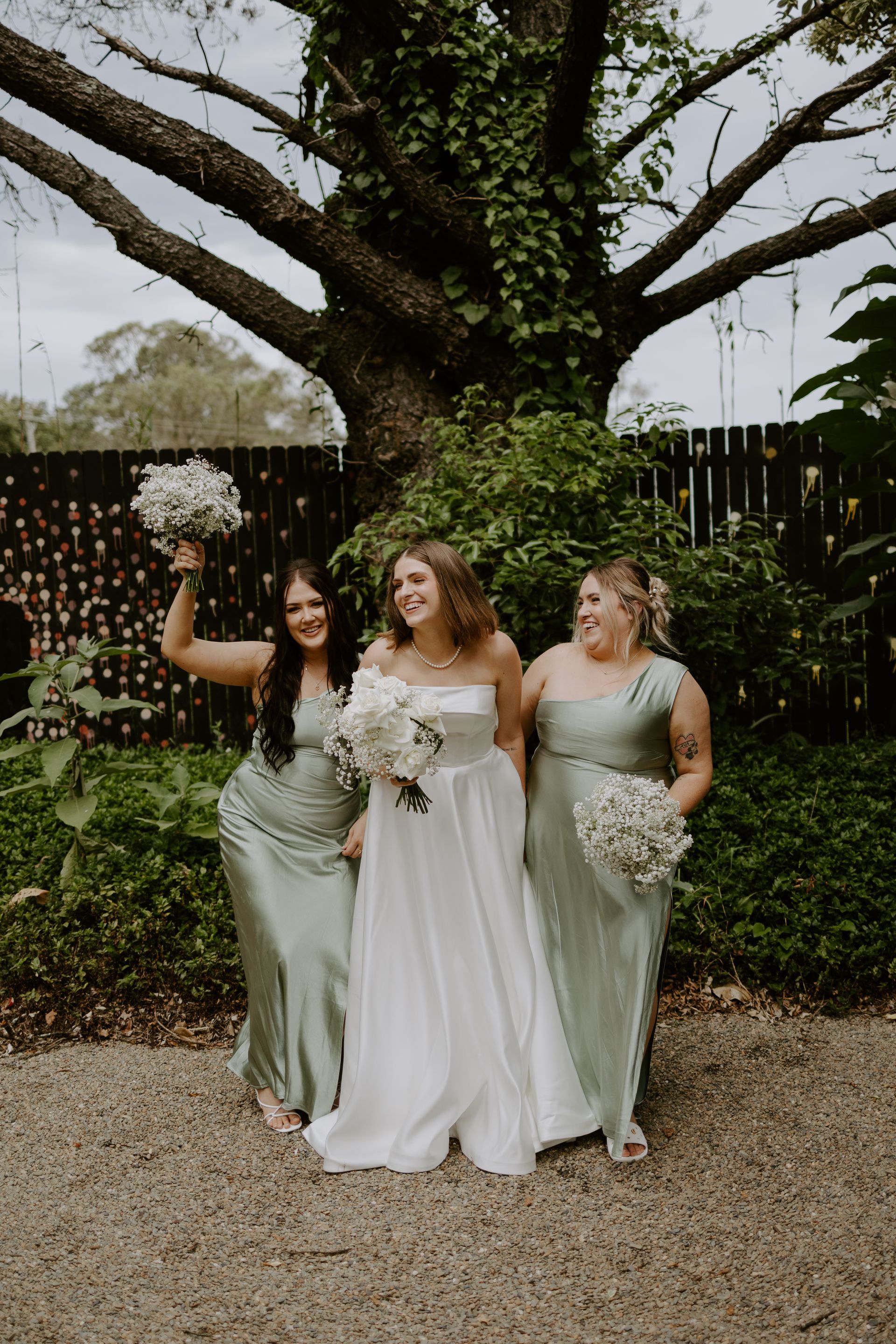 Three bridesmaids in sage dresses and one in white dress posing under a large tree with flower petals falling
