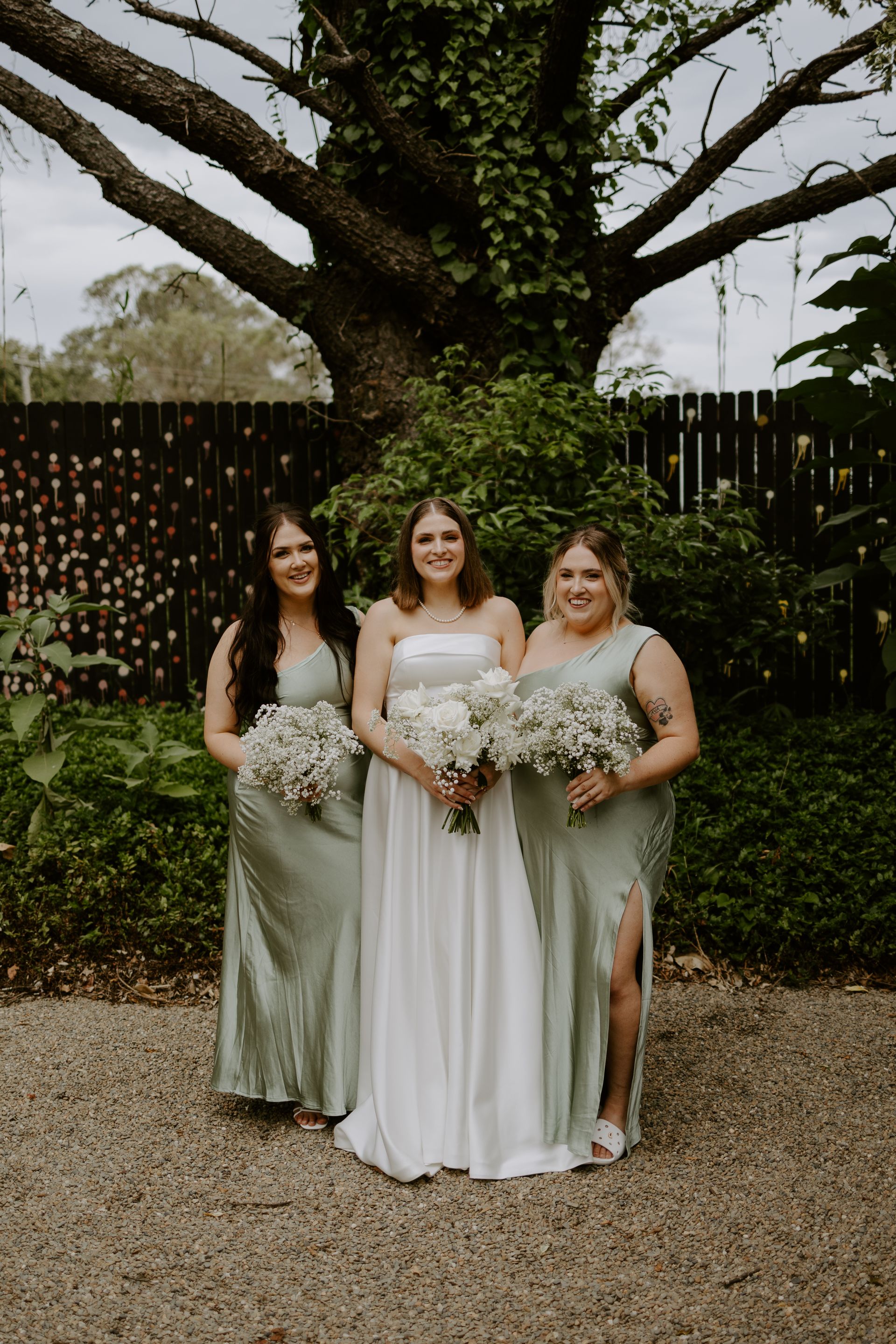 Three women in formal dresses pose outdoors holding white bouquets under a large tree.