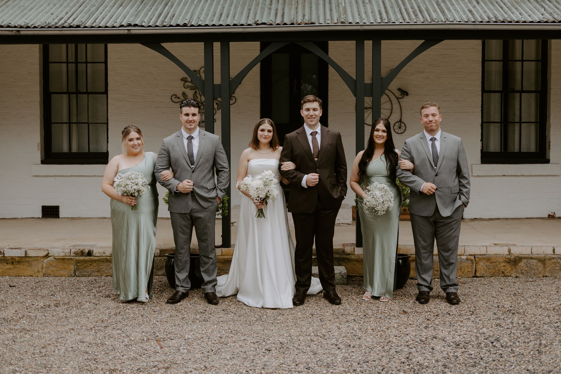Six people in formal attire pose outdoors in front of a rustic building.