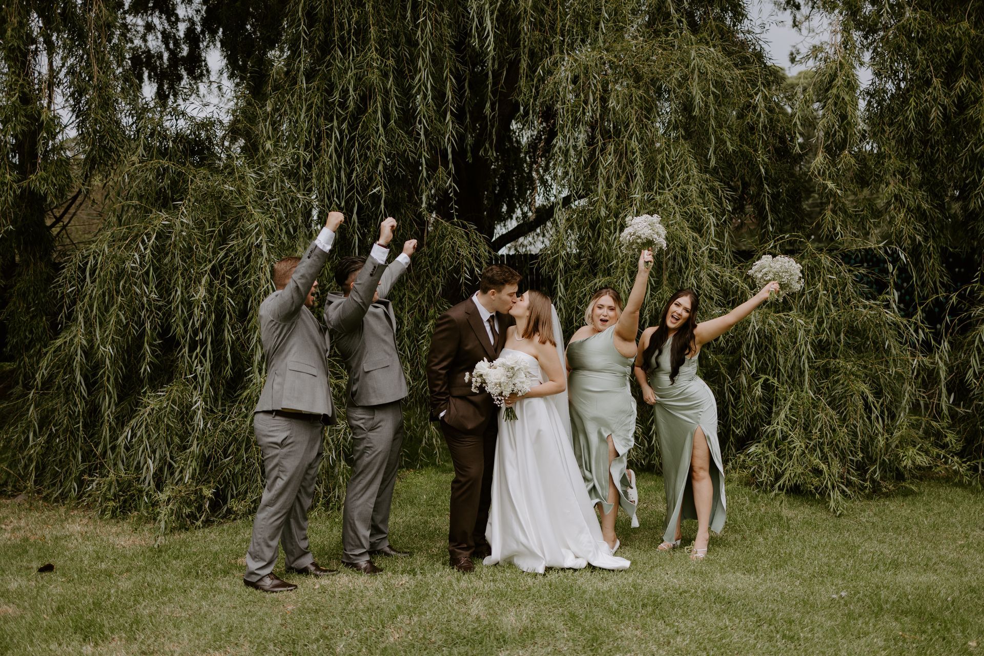 Wedding party celebrating outdoors in a grassy field, standing before a tree with arms raised and bouquets.