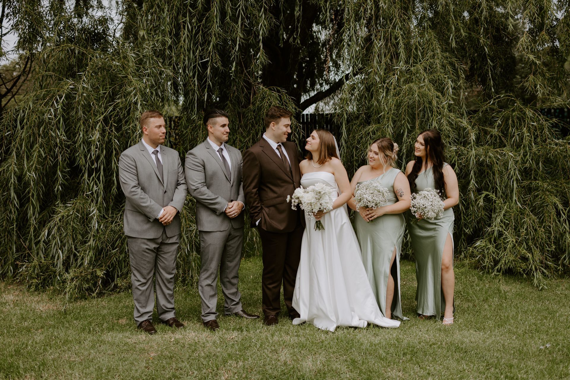 Wedding party standing outdoors in front of leafy trees, with the bride and groom centered.