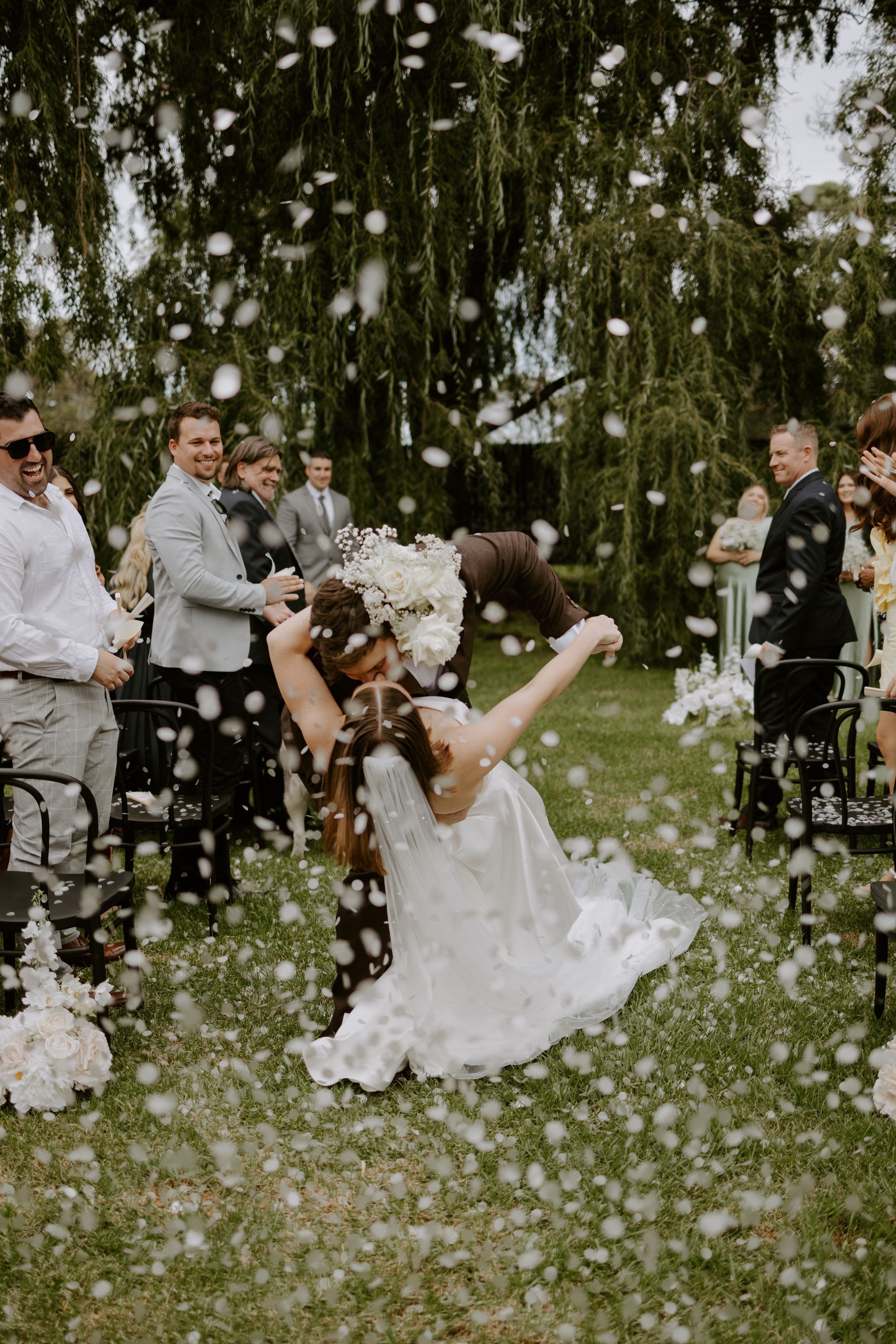 Wedding couple caught in a burst of white confetti outdoors, guests smiling nearby