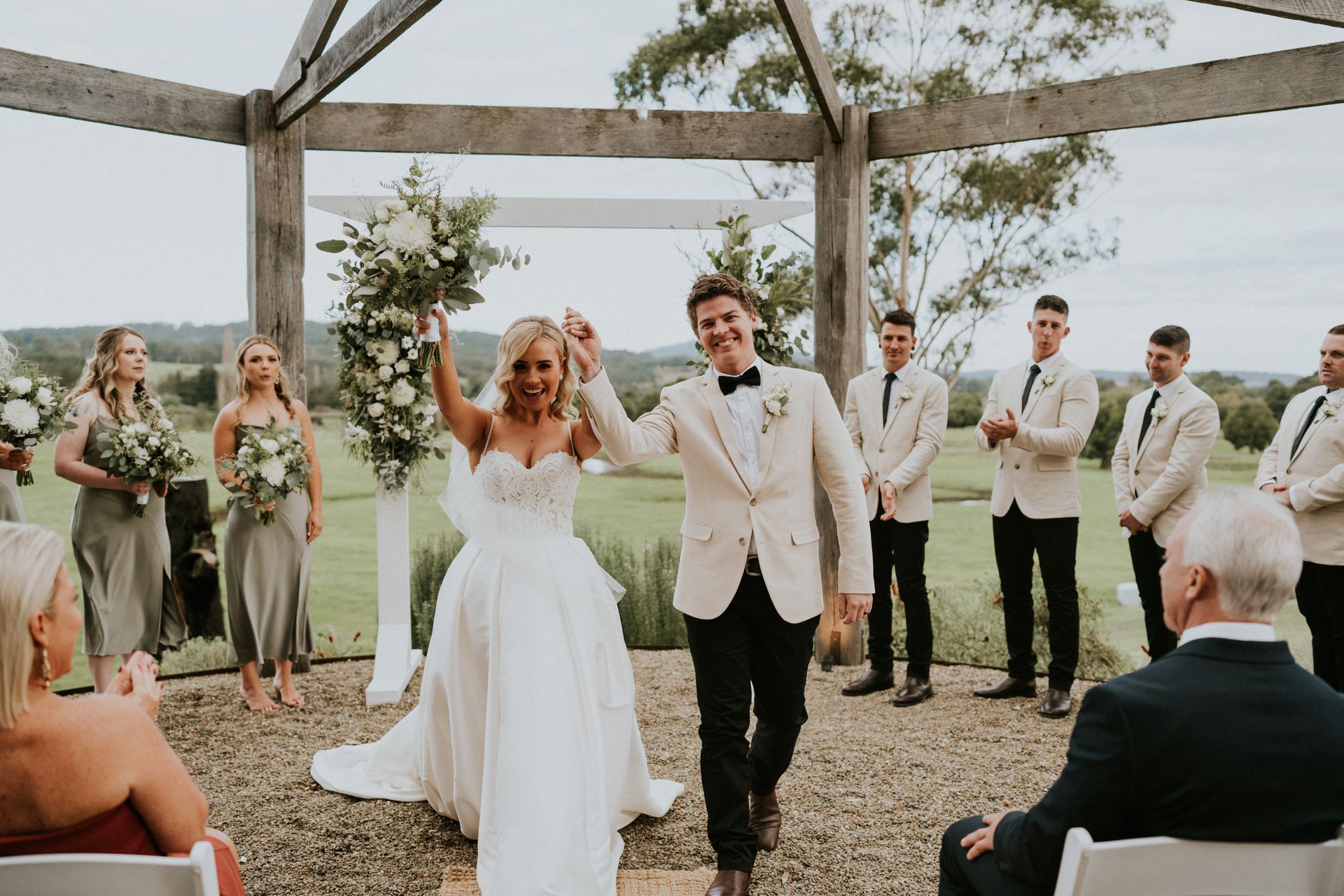 Bride and groom walk down the aisle after the ceremony, guests applauding under a rustic outdoor pavilion.