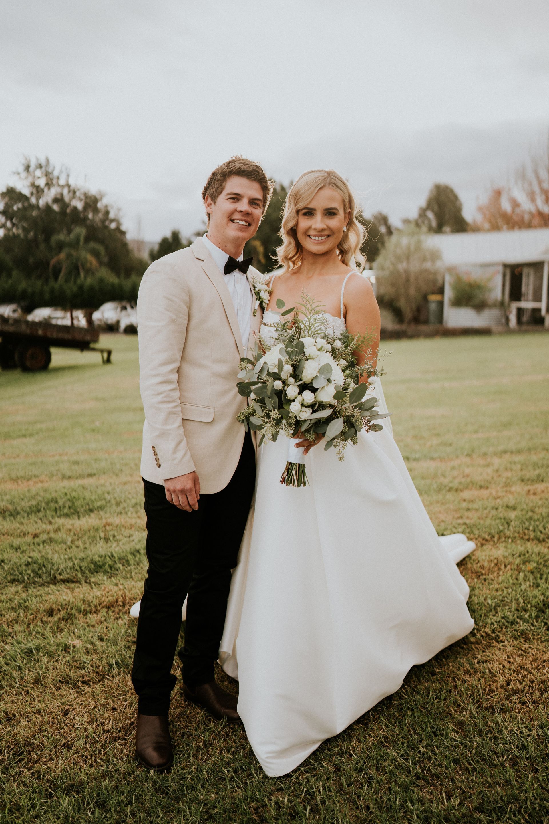 Two people posing outdoors at a wedding, holding a bouquet, with a grassy field and building in the background.