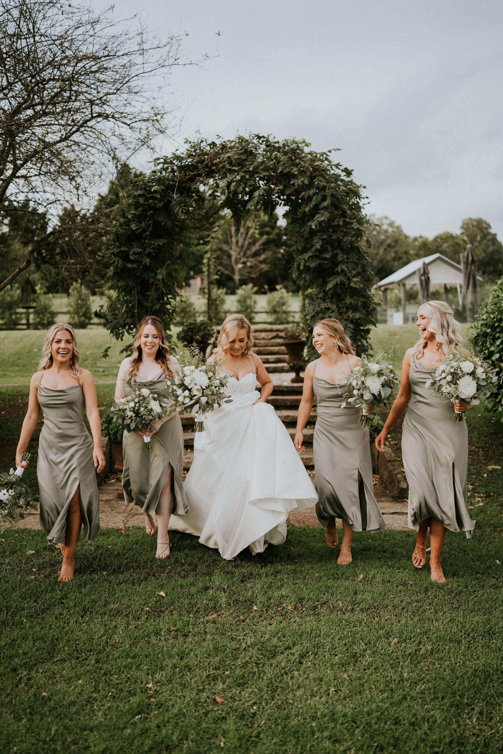 Bride and bridesmaids posing outdoors in white and sage dresses in front of a floral arch