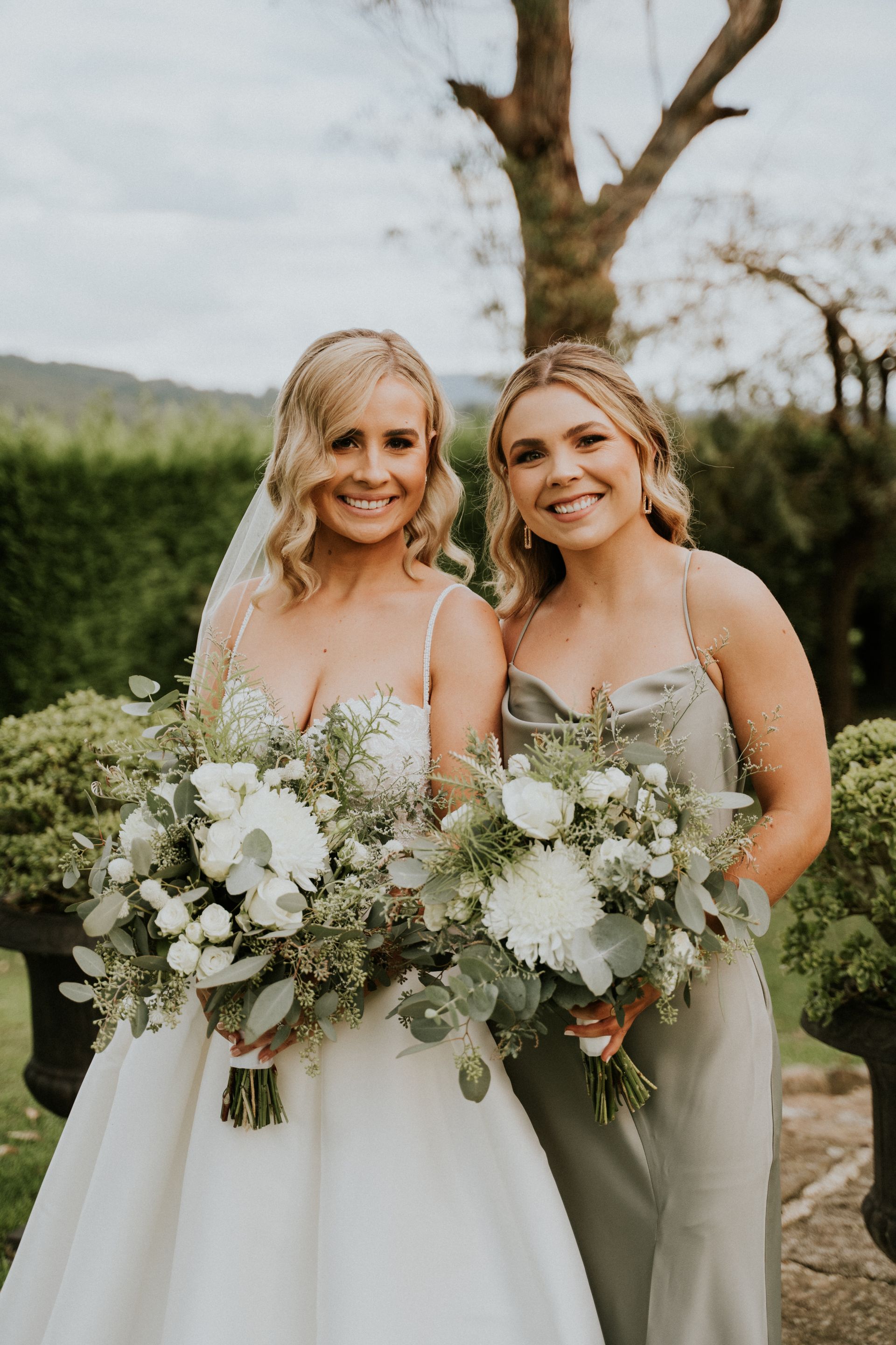 Two brides in white dresses holding large white bouquets outdoors, smiling beside a tree.