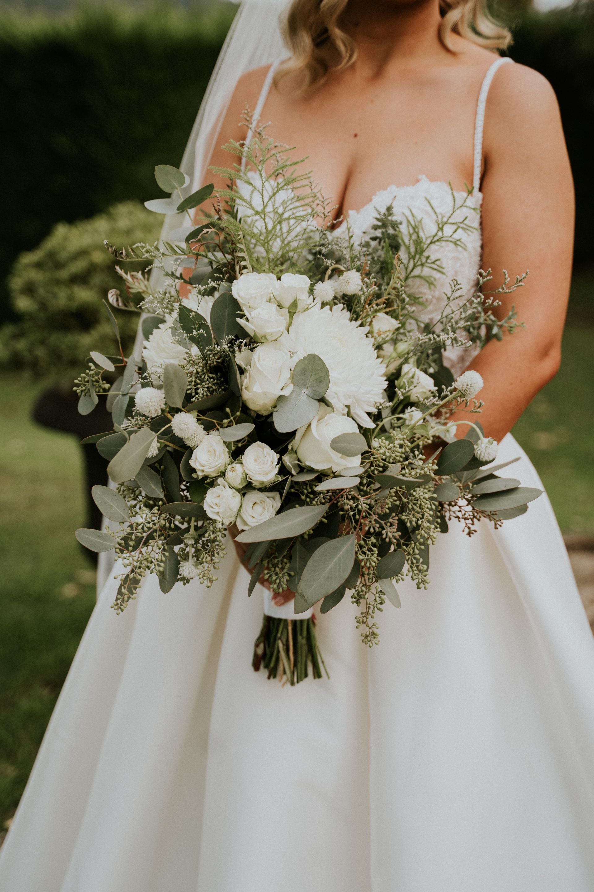 Bride in a white dress holding a lush white-and-green bouquet outdoors