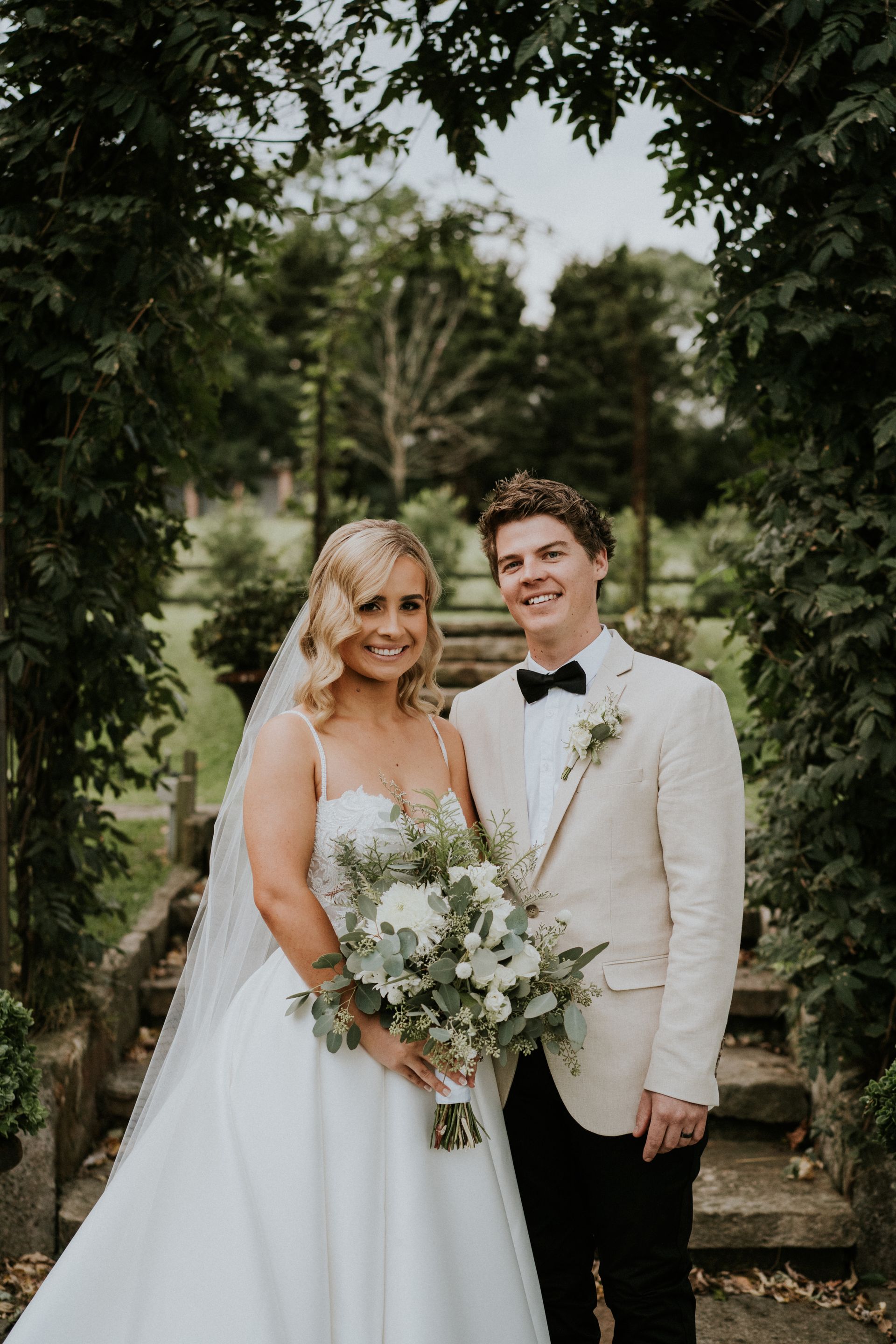 Bride and groom posing outdoors in wedding attire, holding a bouquet under greenery.