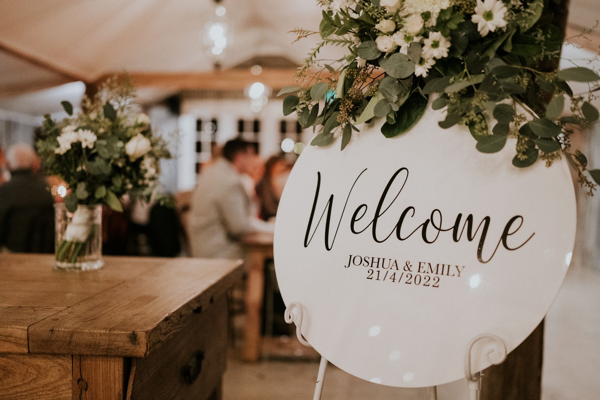 Rustic wedding welcome sign on a wooden post with white flowers and blurred reception guests in the background
