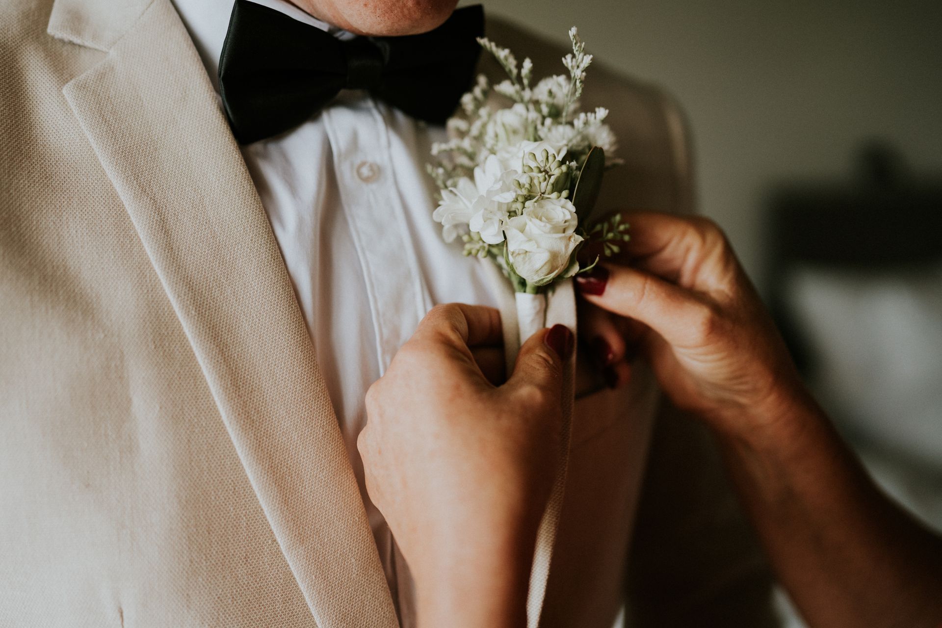 Hands pinning a white boutonniere onto a beige suit jacket and bow tie