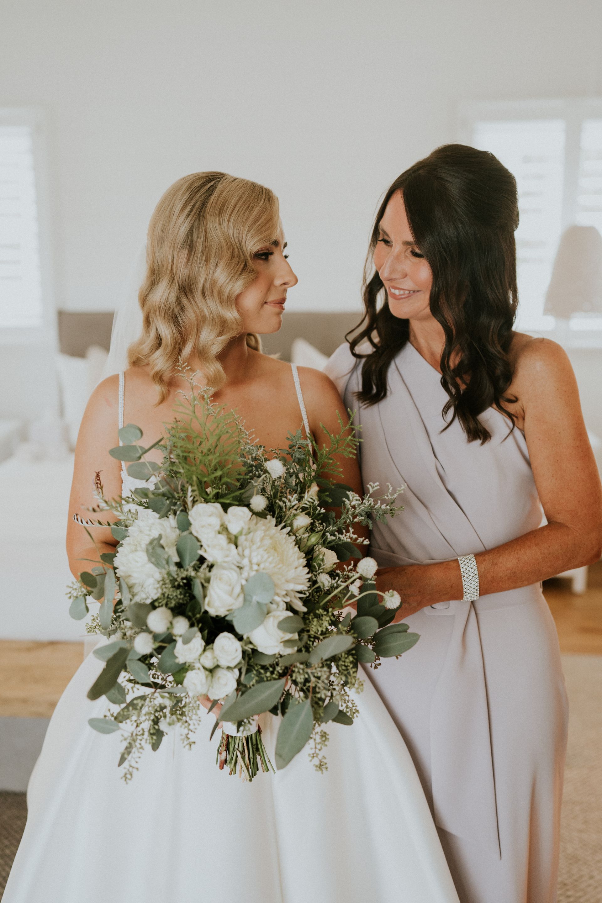 Two women in white dresses smiling, one holding a large white-and-green bouquet indoors.