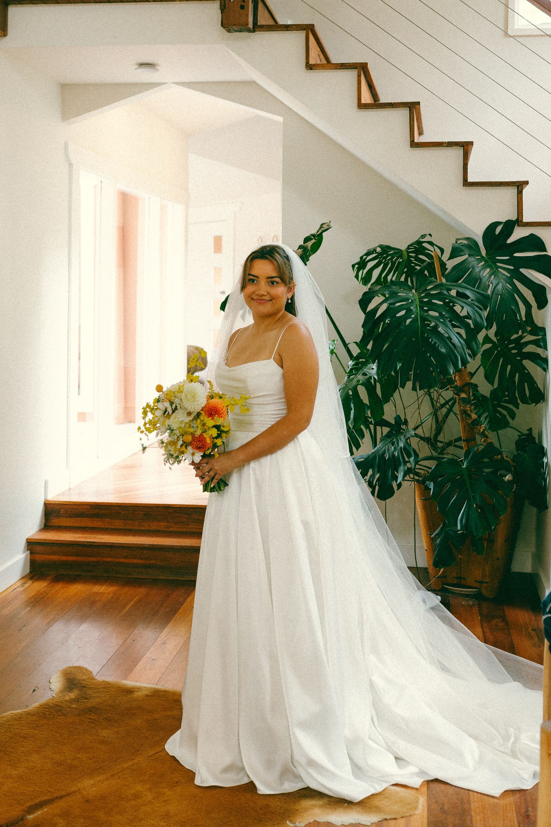 Bride in a white wedding gown holding a bouquet, standing indoors by a staircase and potted plant.