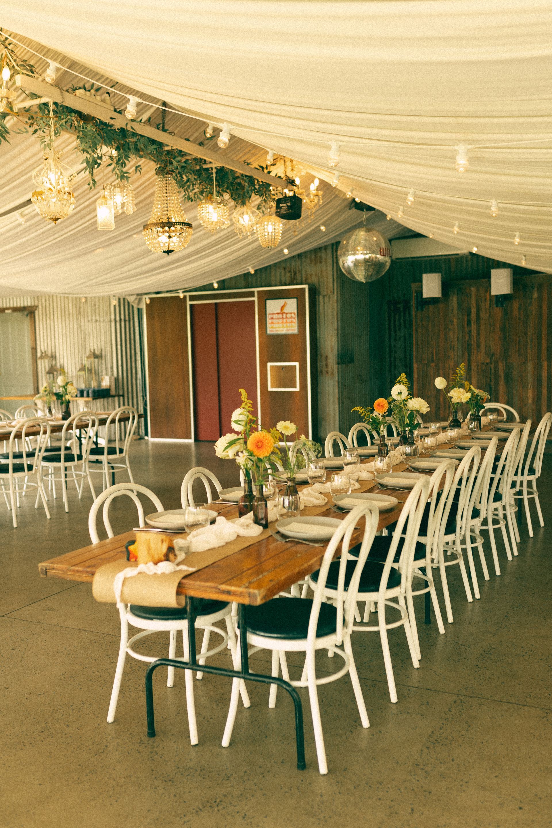 Decorated banquet hall with long table, white chairs, floral centerpieces, and draped ceiling lights