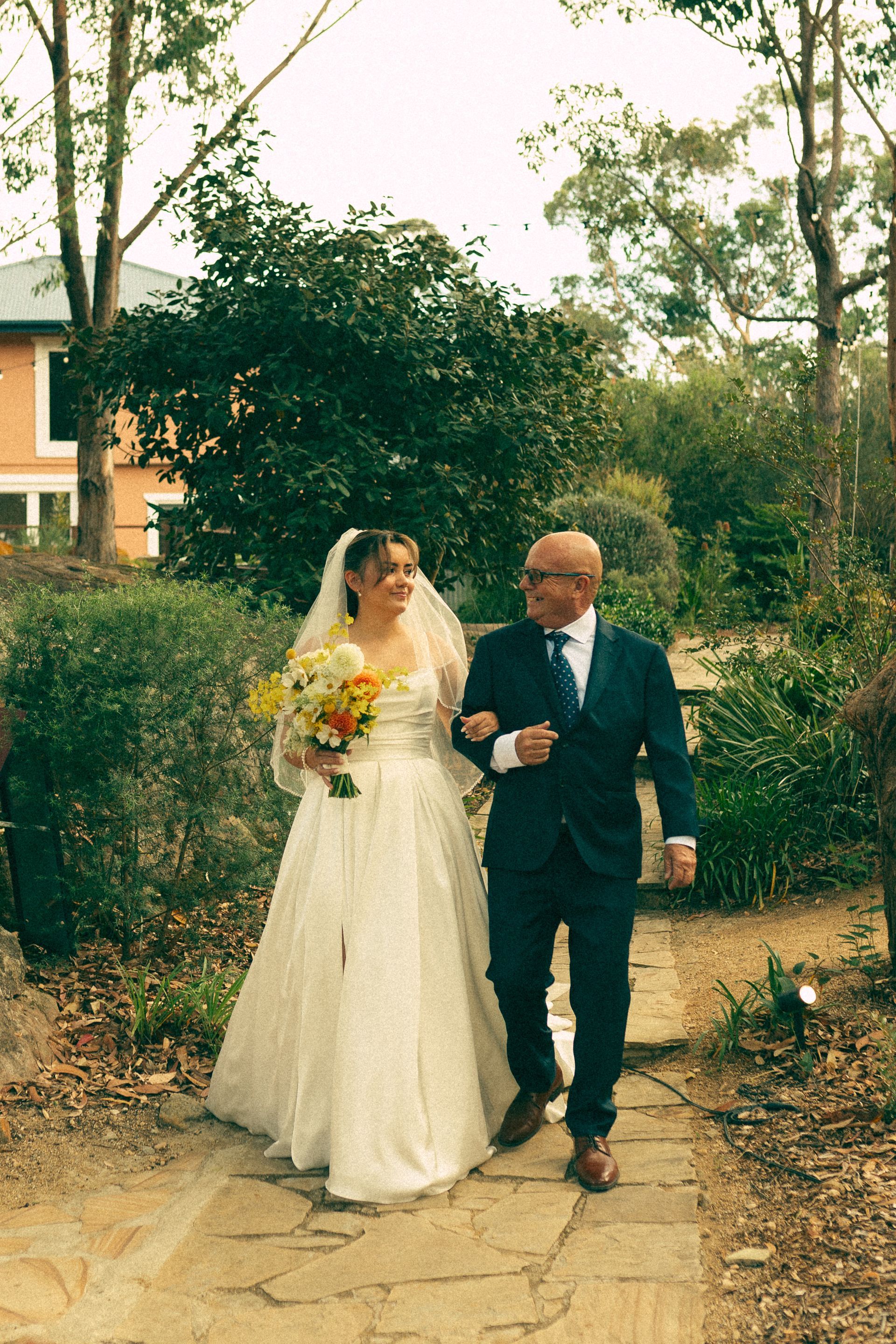 Bride in white dress walking with a man in a dark suit along a garden path.