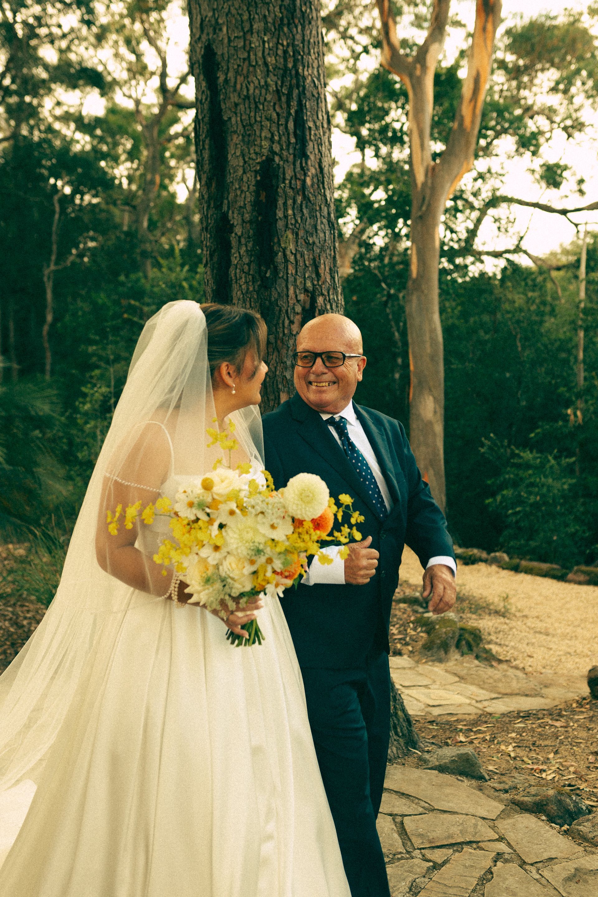 Bride in white gown holding bouquet walks with suited companion outdoors on a garden path.