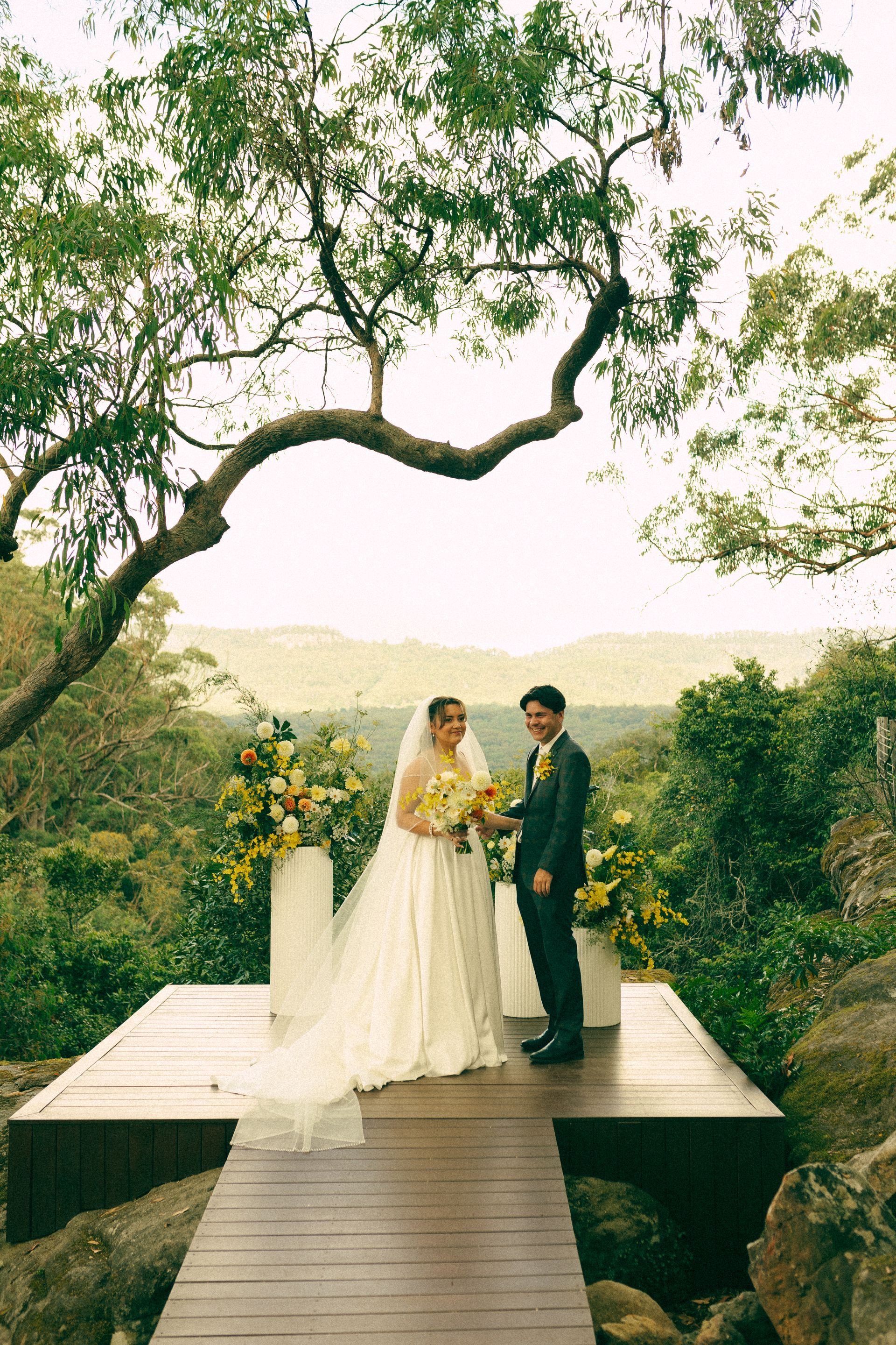 Wedding couple standing on a wooden platform under a large tree, surrounded by greenery and flowers