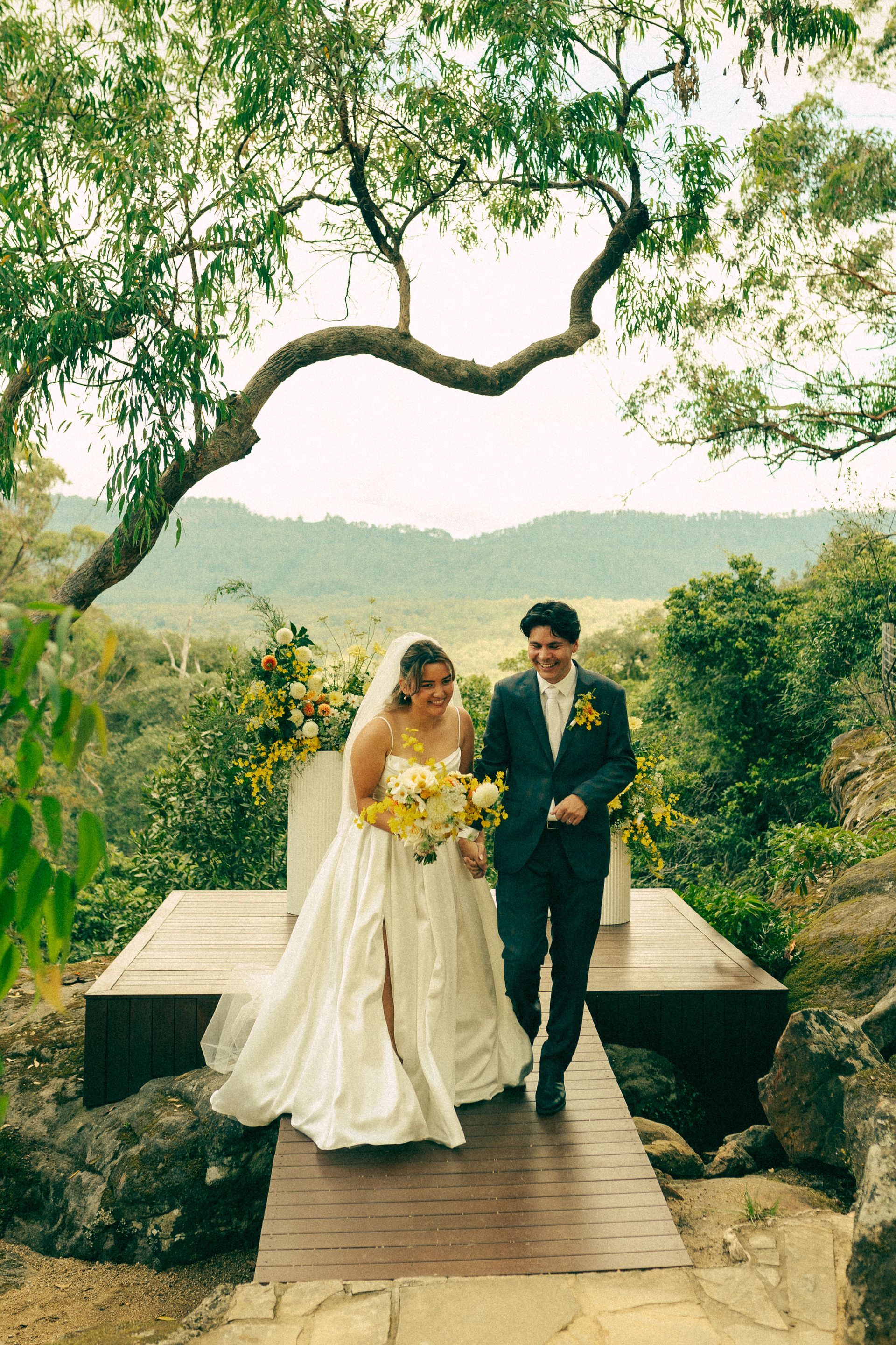 Bride and groom walking on a wooden bridge in a lush garden, with mountains in the background