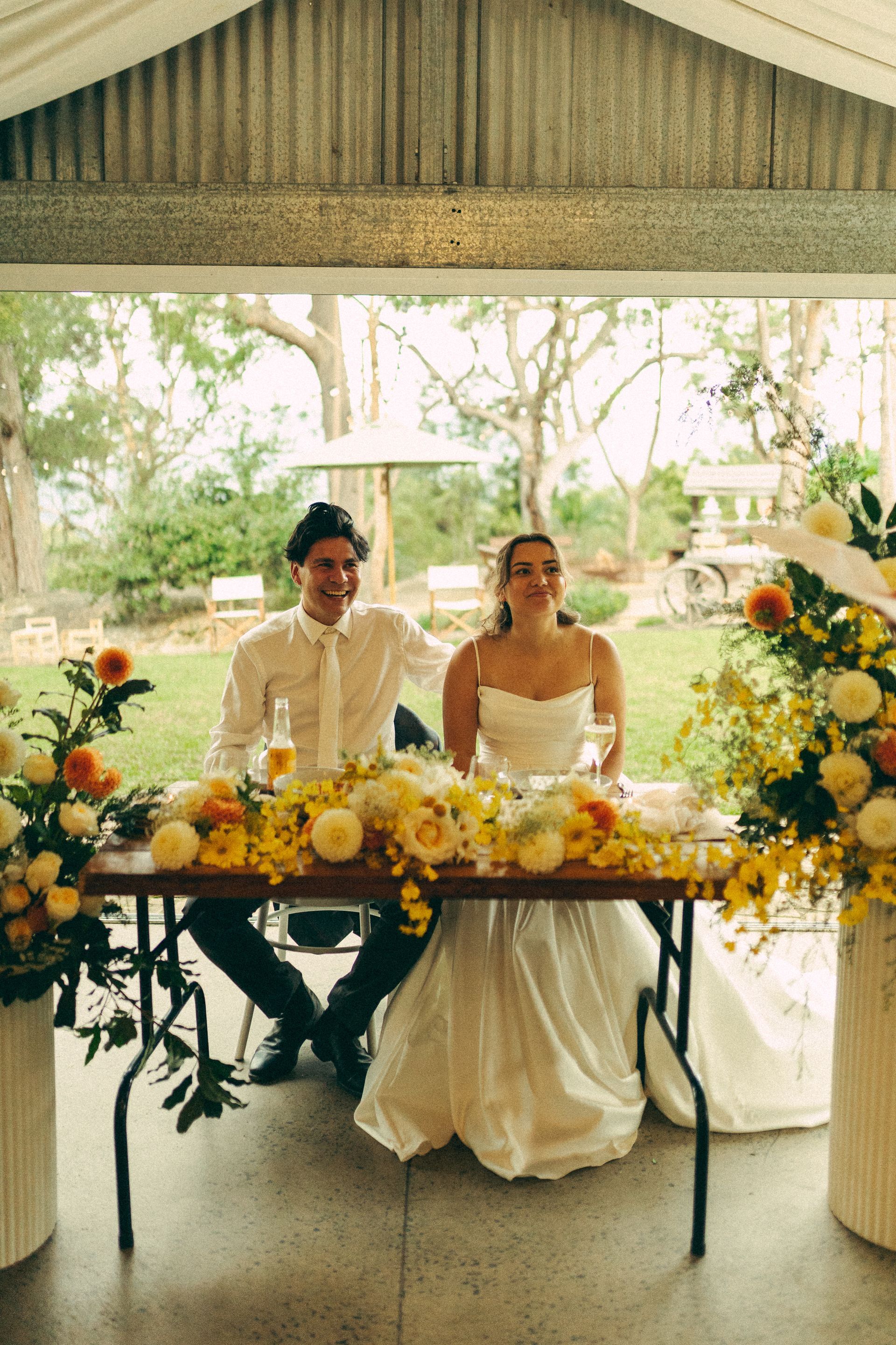 Two people seated at a flower-decorated wedding table in a bright tent, smiling at the camera